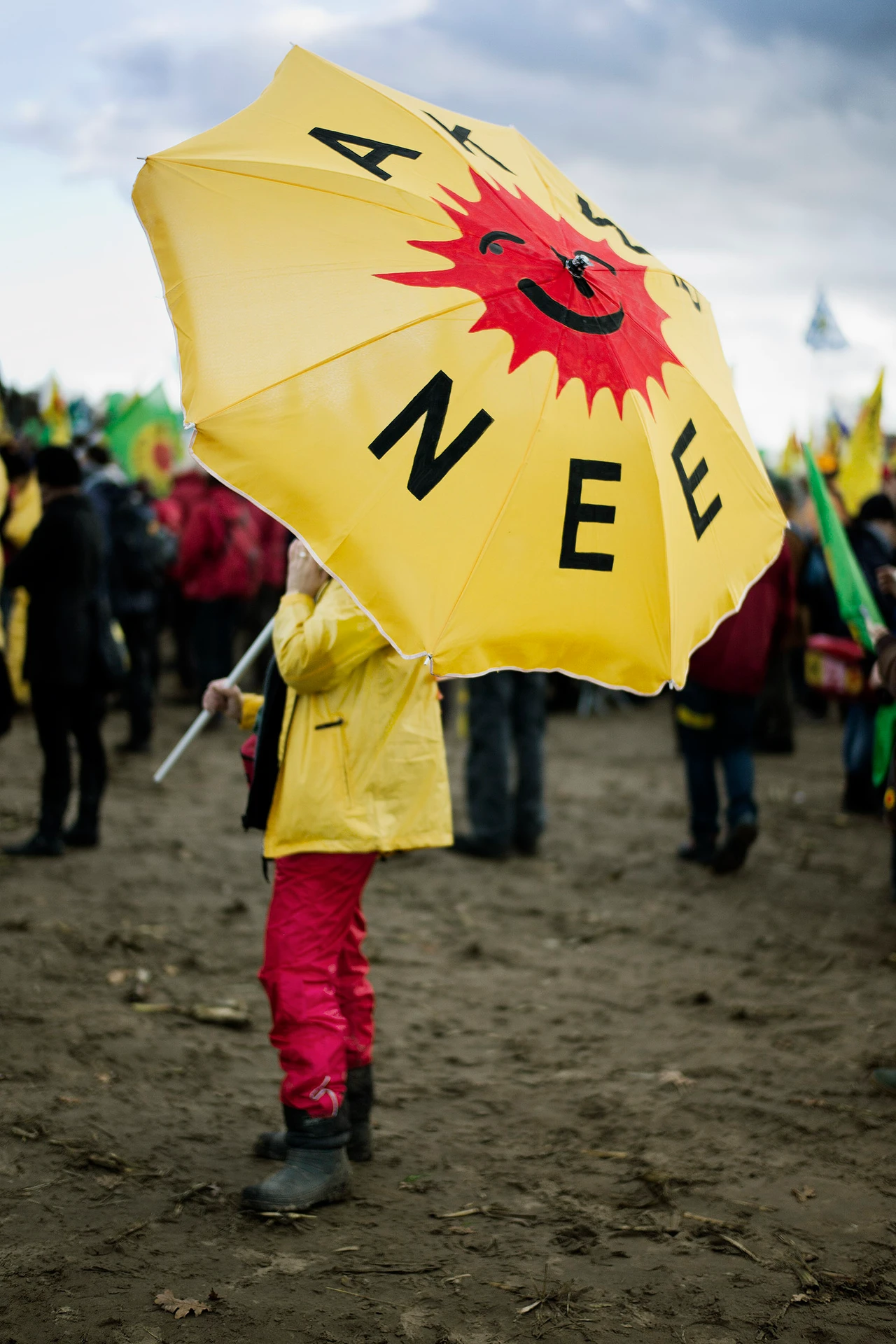 A person wearing a yellow raincoat and red trousers holds a yellow parasol bearing the inscription AKW NEE (no nuclear power plants) and a smiling red sun on a muddy protest site.