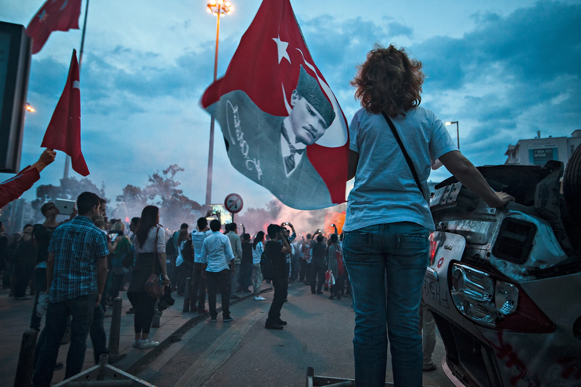 A person holding an Atatürk flag photographed from behind stands next to a damaged car at night and looks at a large crowd of people. Thick smoke and several demonstrators can be seen in the background.