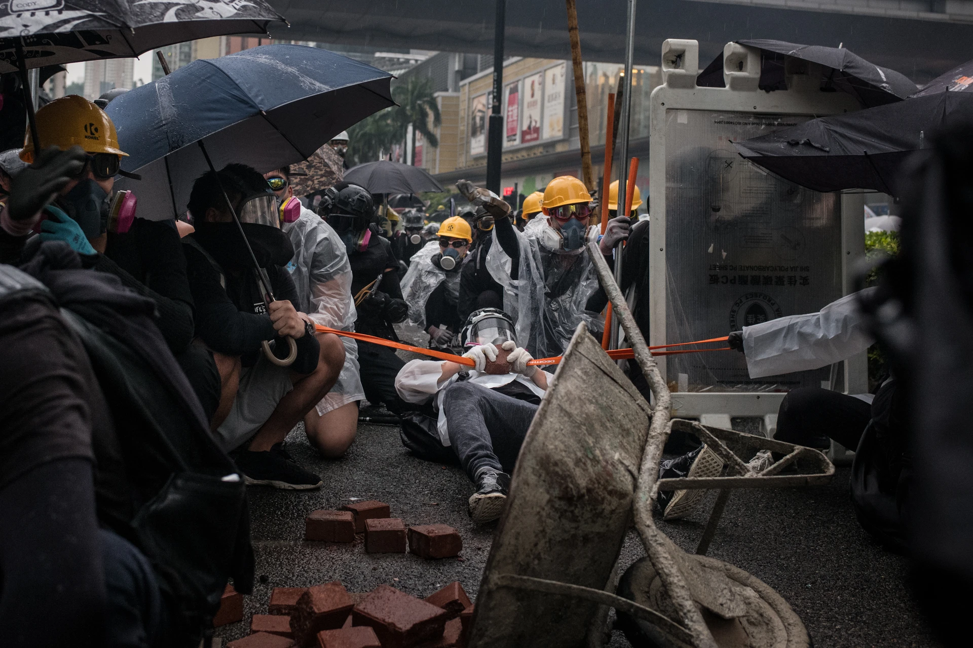 On a street in Hong Kong, several masked people wearing hard hats and rain gear hurl a slingshot behind an improvised barricade.