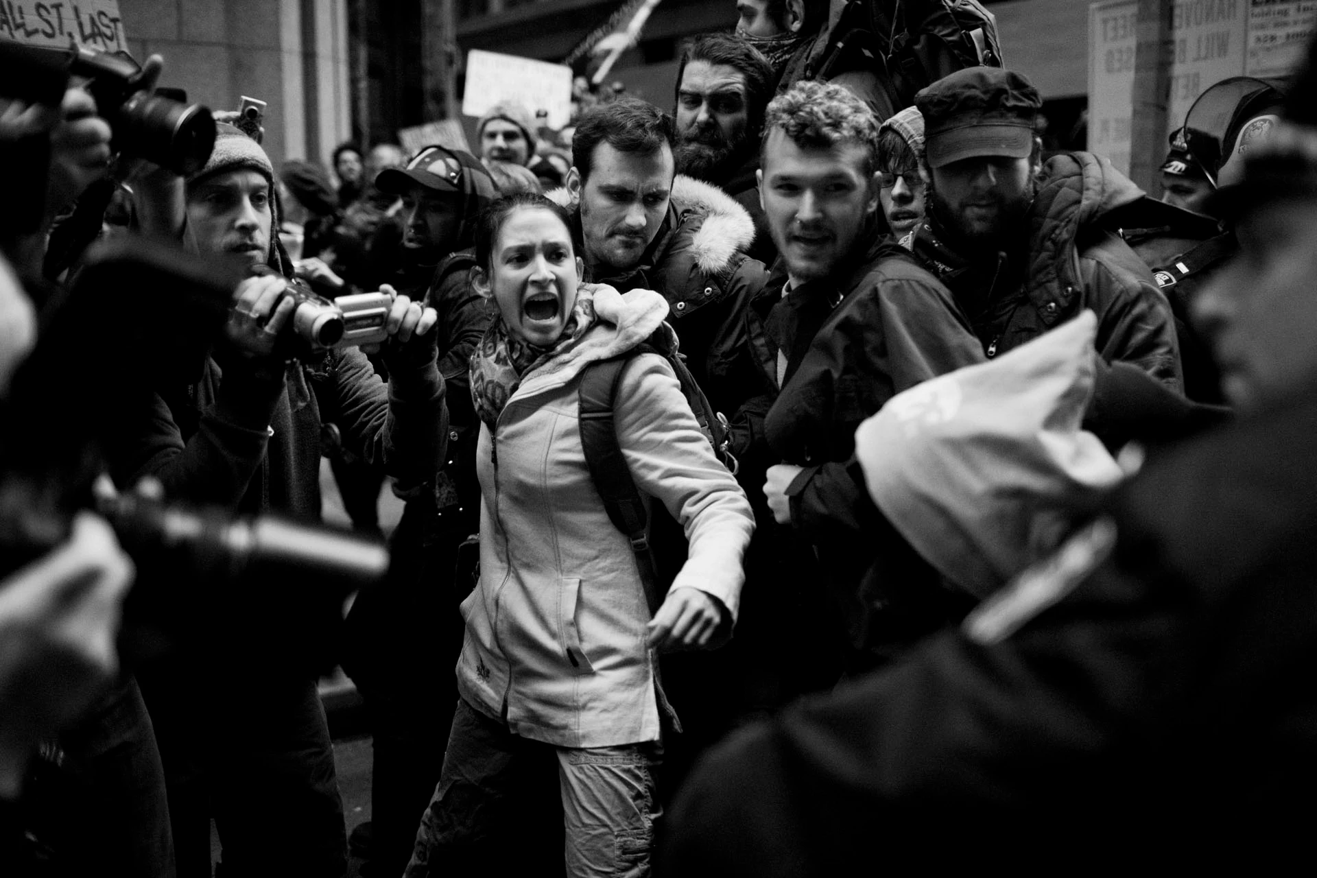 Black-and-white image: A young woman surrounded and held by several people, some of whom are holding cameras, shouts at a police officer in the foreground.