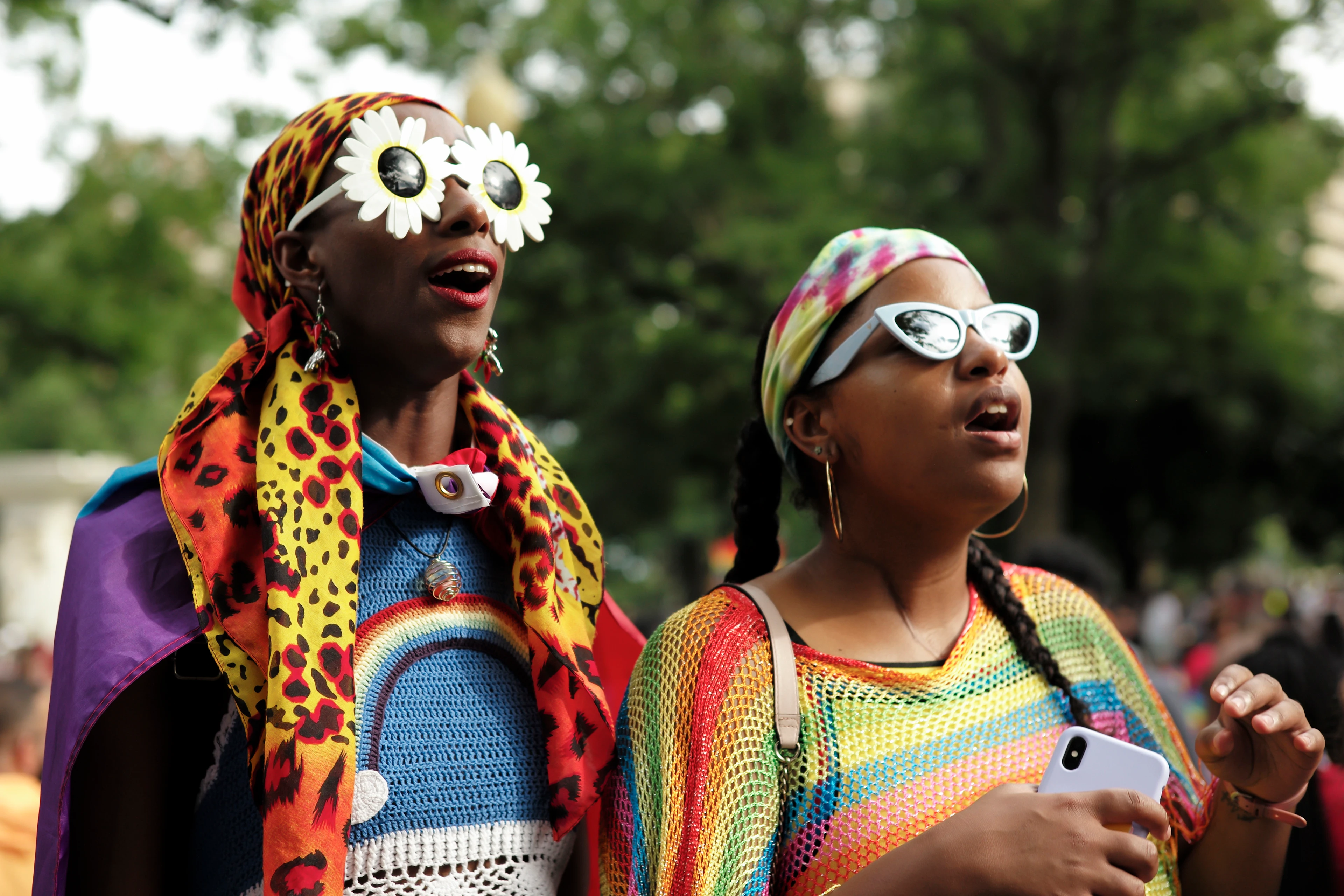 In this image, two Black women wearing colourful clothing and eye-catching sunglasses stand side by side, smiling with slightly open mouths as they take part in an outdoor parade.