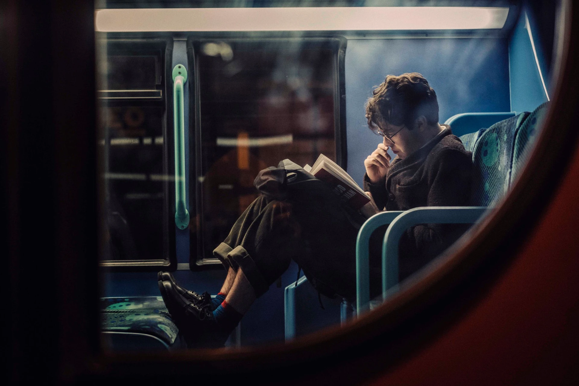 An atmospheric view of a man engrossed in a book with his knees drawn up, as seen through a bus window.