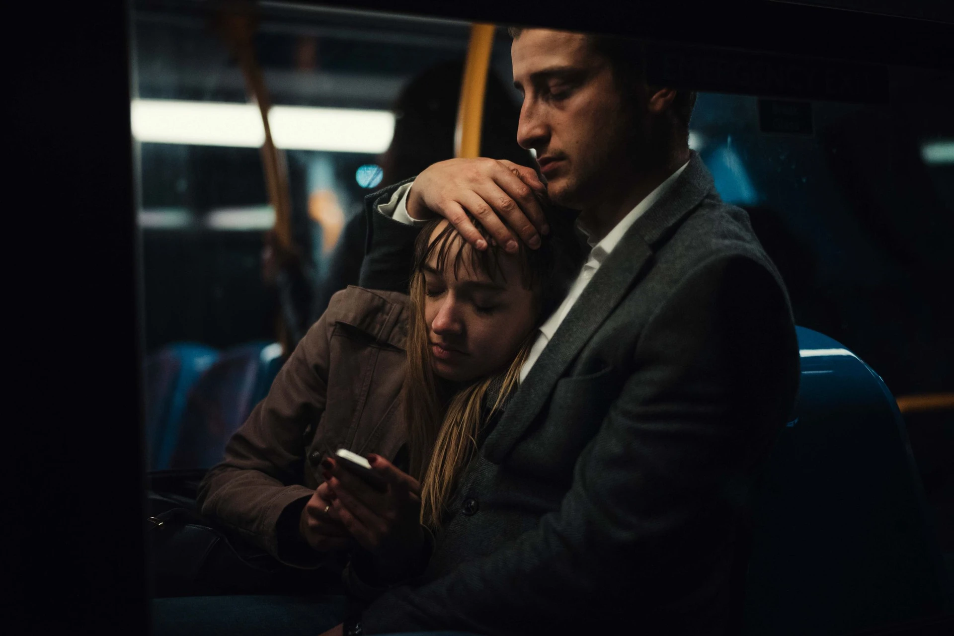 A couple sit close together by the bus window at night. He holds his hand on her head with his eyes closed while she looks at her smartphone.