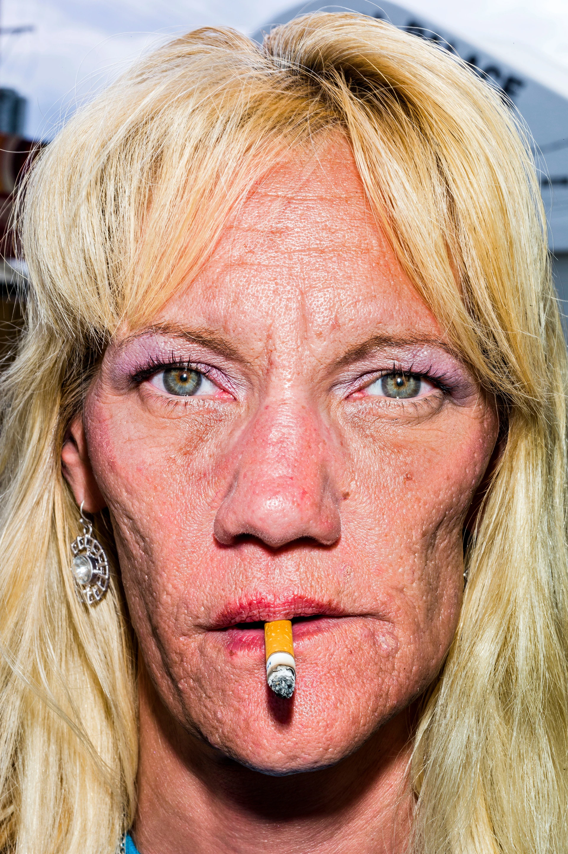 一位染着金发、涂着粉色眼影、嘴里叼着烟头的女子,脸上刻满生活的印记,表情僵硬地直视着相机镜头,闪光灯将她的脸庞照得格外清晰。