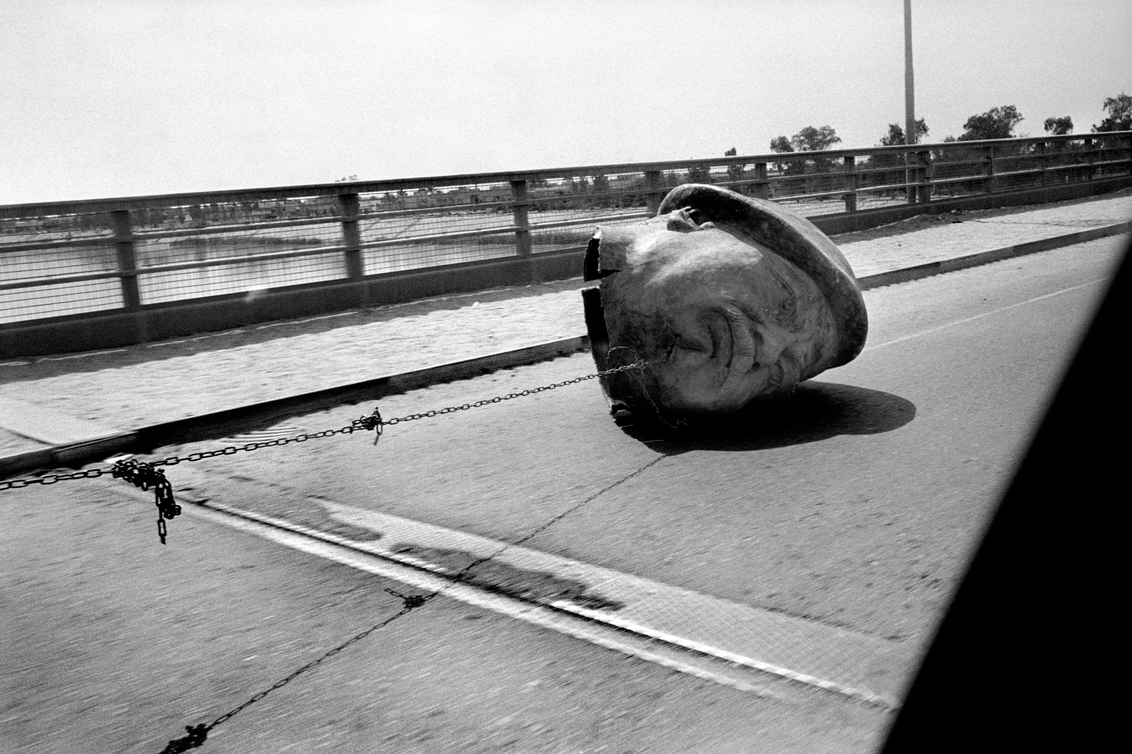 Schwarz-Weiß-Bild vom Kopf einer großen Saddam Hussein Statue die an einer Stahlkette hängend über eine Autobrücke gezogen wird.