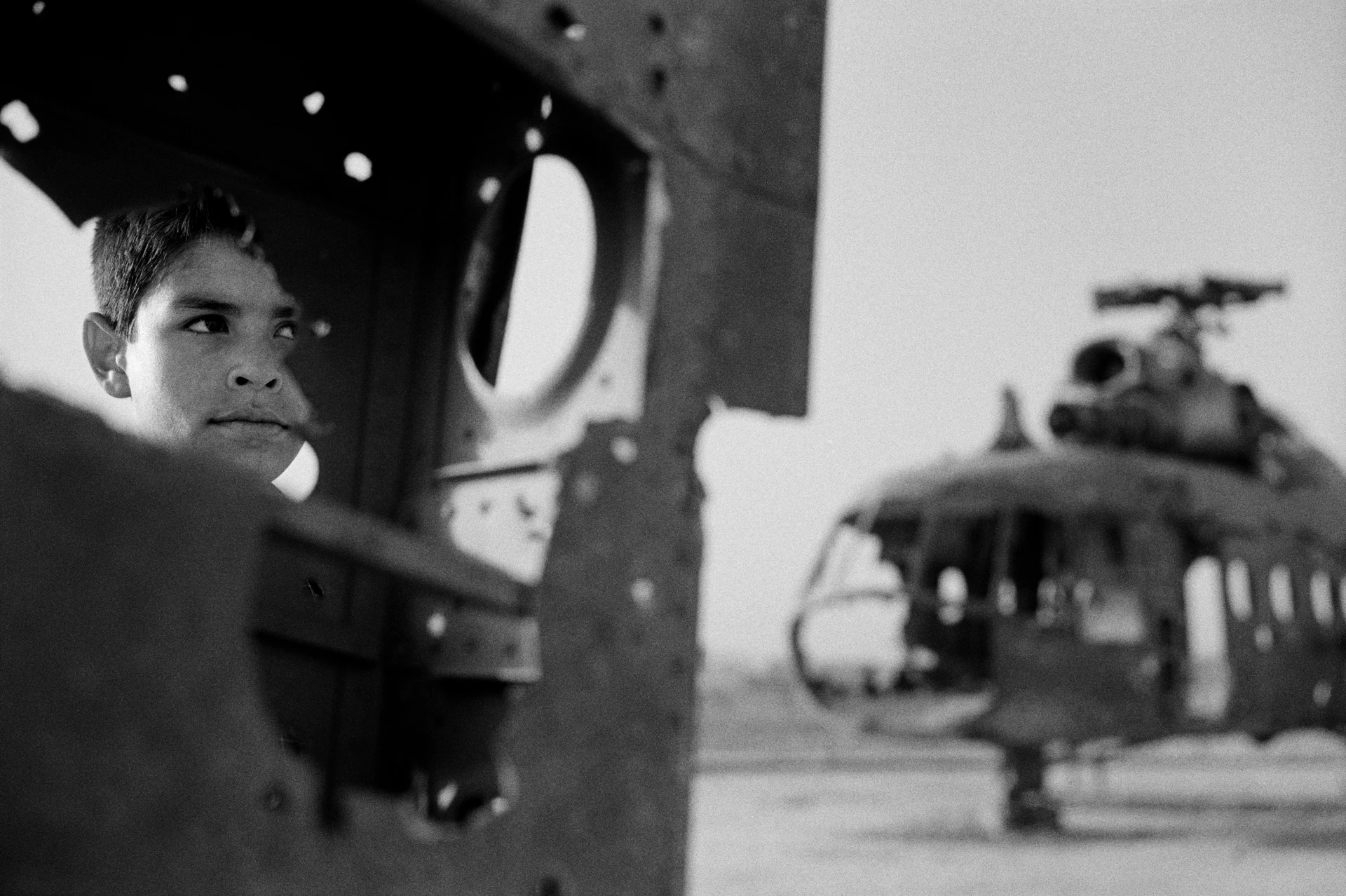 Photographie dramatique en noir et blanc d’un garçon au regard fixe photographié à travers un trou dans une épave de char, les restes d’un hélicoptère se trouvent à l’arrière-plan.