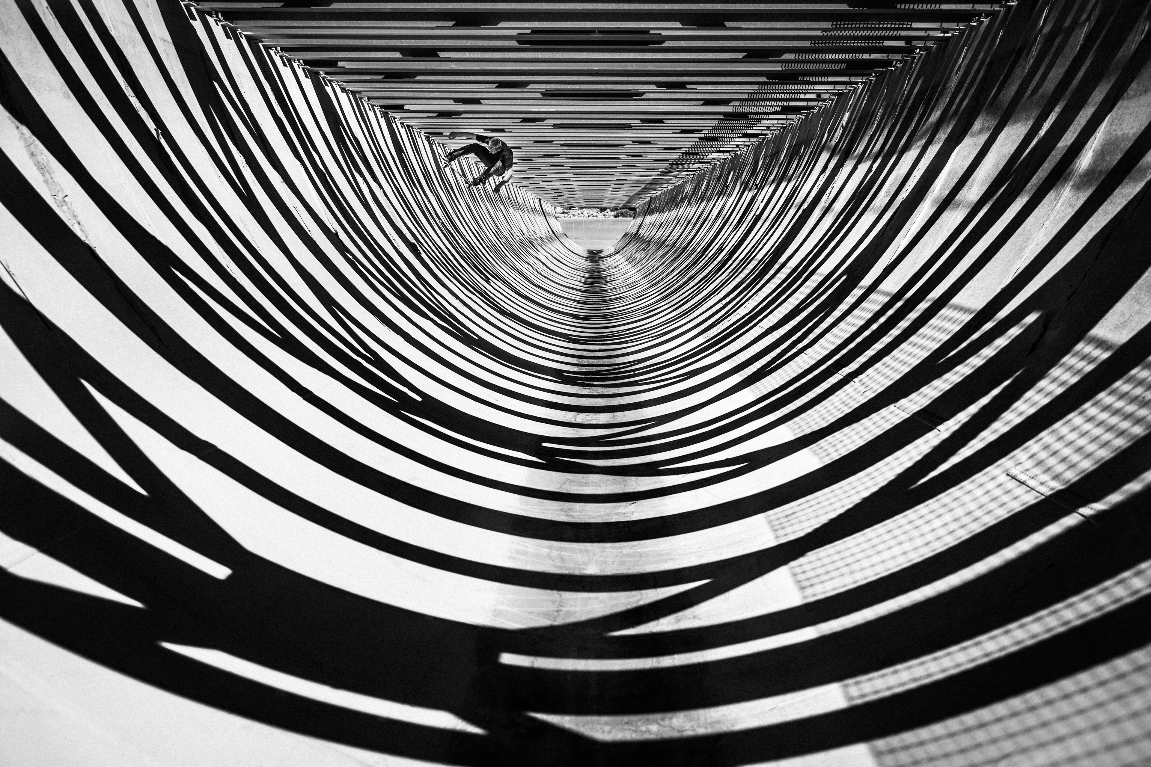 Graphic black-and-white image: A skateboarder rides in a deep, tubular concrete channel. The struts above cast rhythmic, arcing shadows, creating a captivating structure.