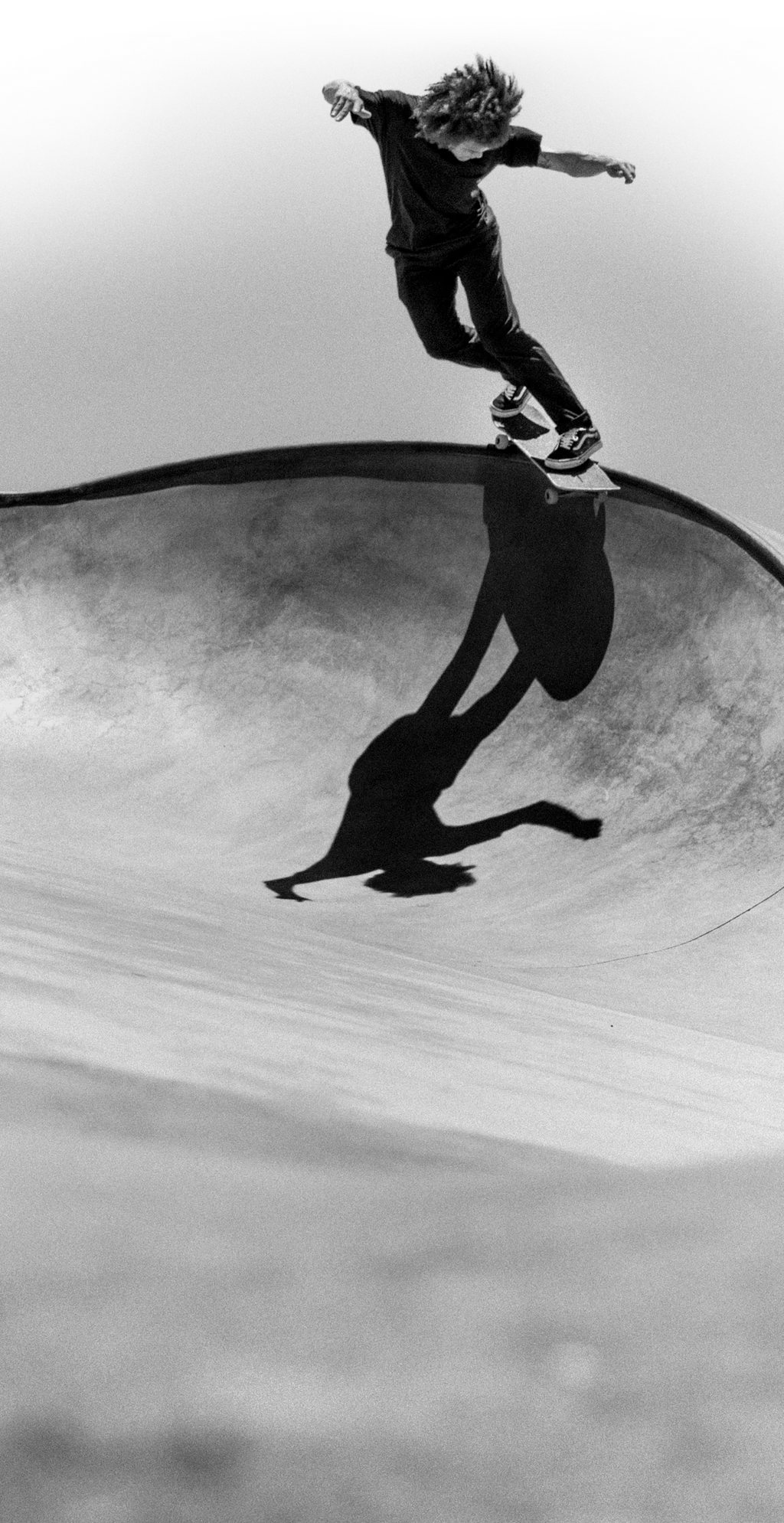 Elegant black-and-white image: A skater glides on the edge of a curved bowl, seemingly weightless against the backdrop of an empty, glistening sky.