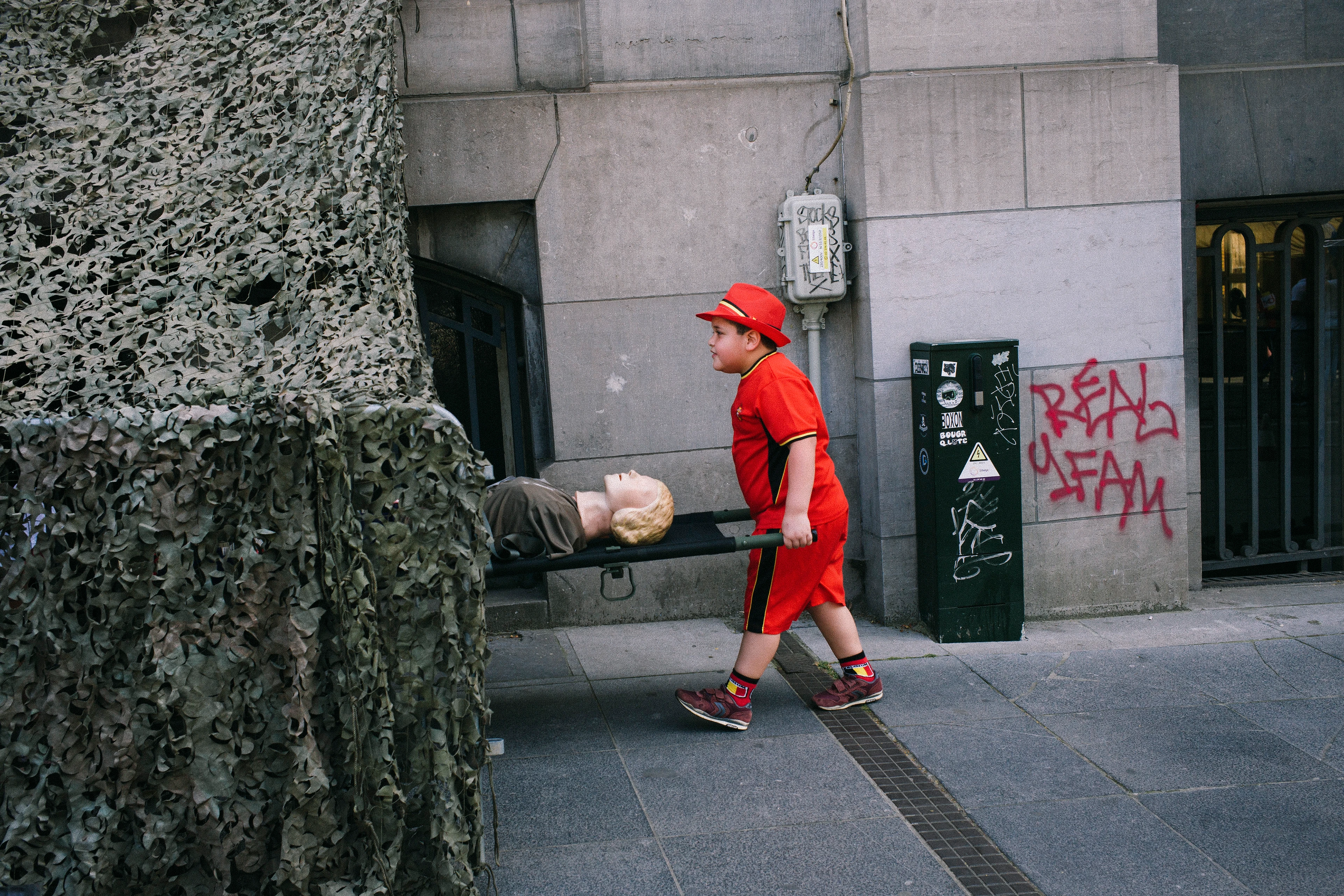 A boy wearing red clothes and a red hat pushes a mannequin on a flatbed trolley into a structure covered with camouflage netting.