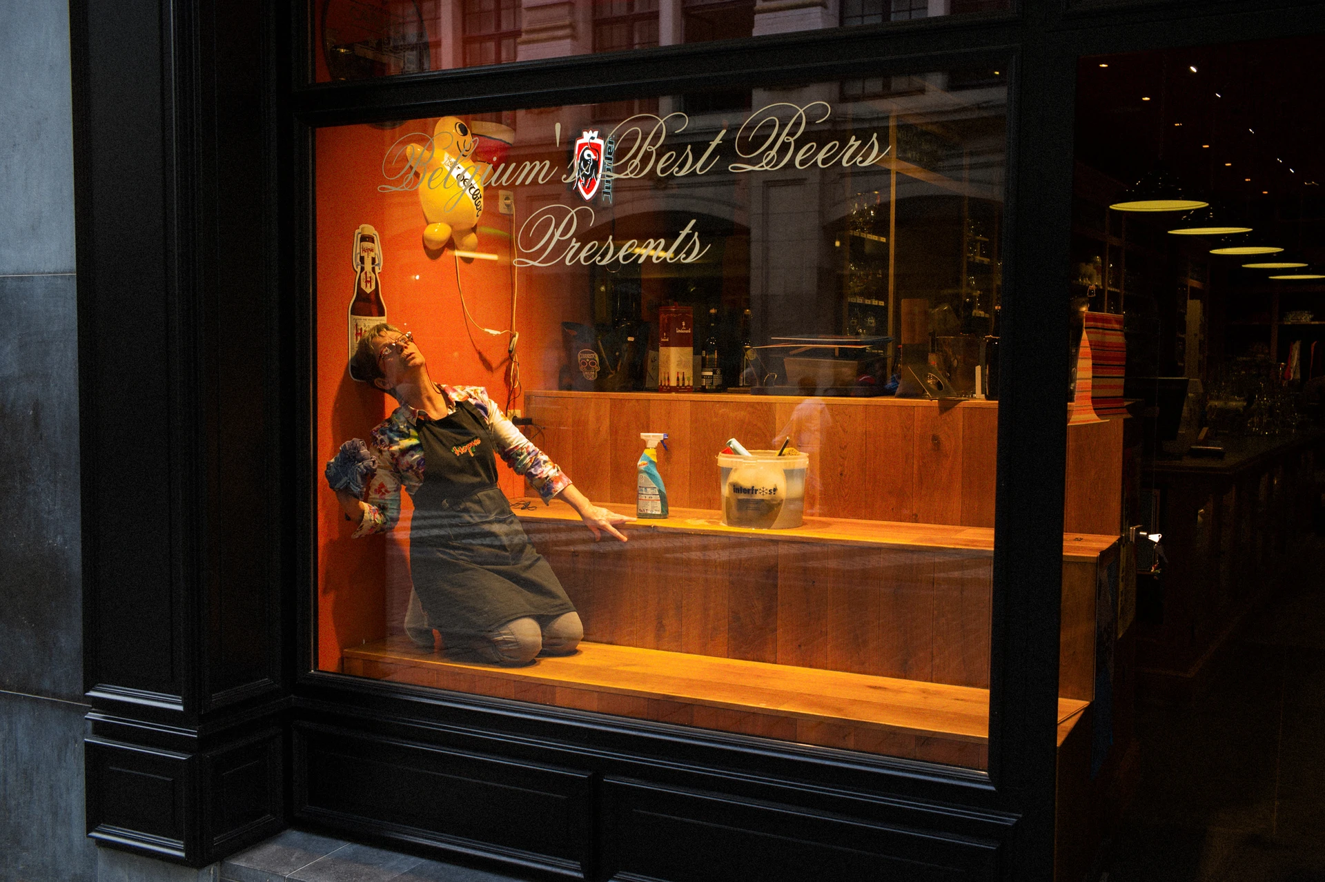 An illuminated orange window display shows a woman cleaning while looking up, like a mannequin.