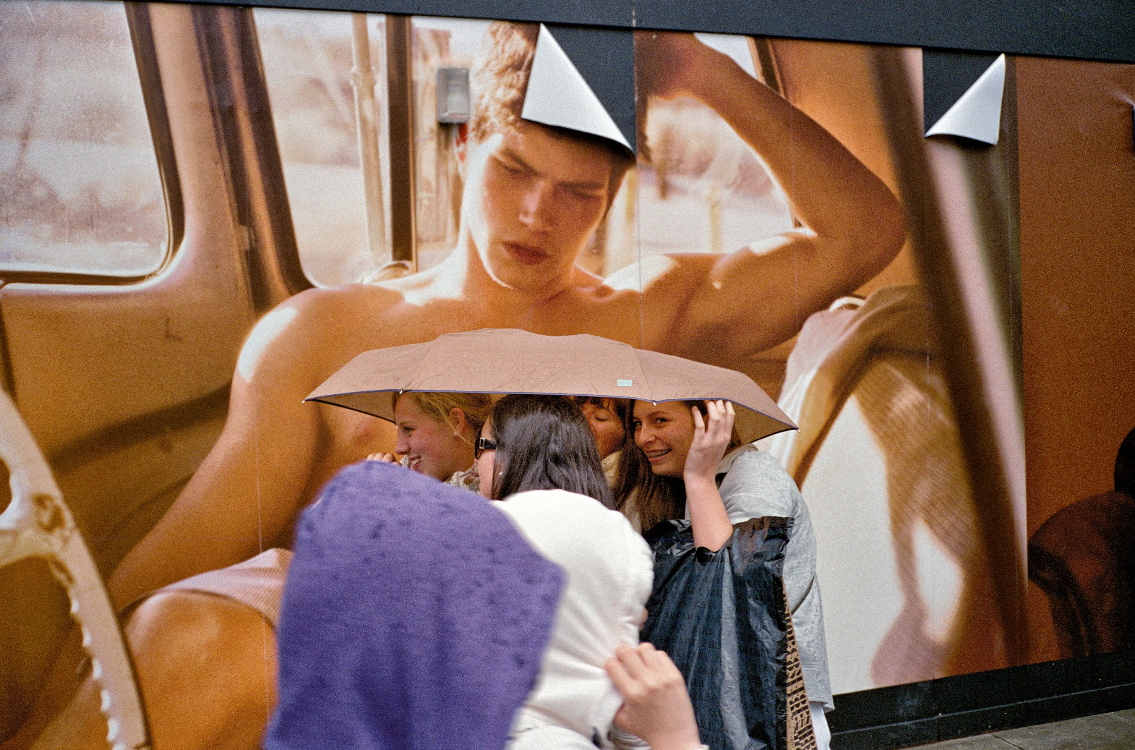 Several women stand smiling together under a beige umbrella. Behind them, a billboard shows a topless young man lasciviously posing and looking down at the ladies.
