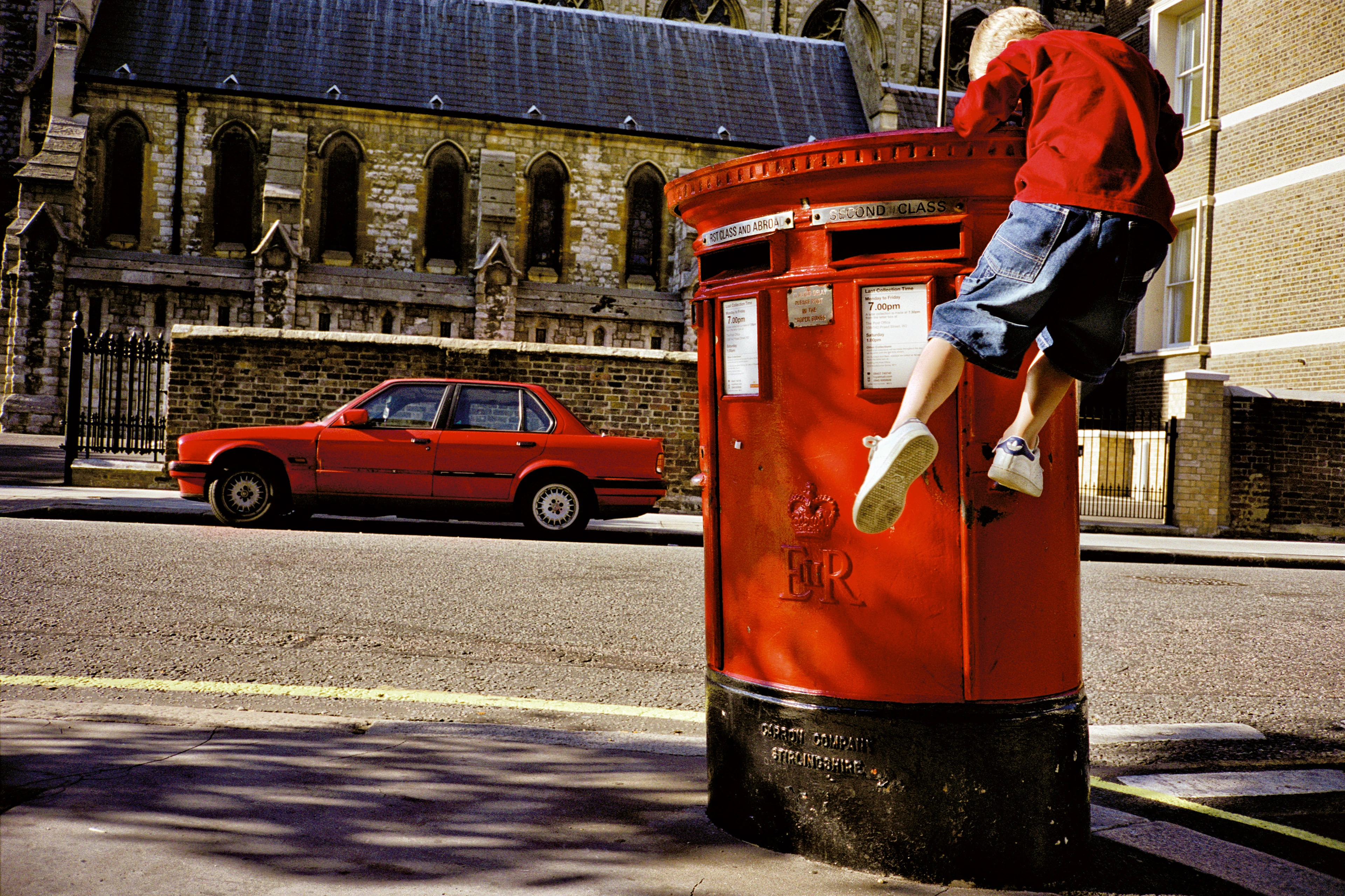 A boy wearing a red jumper climbs a large red public letterbox while a red parked car and an old church building can be seen in the background.