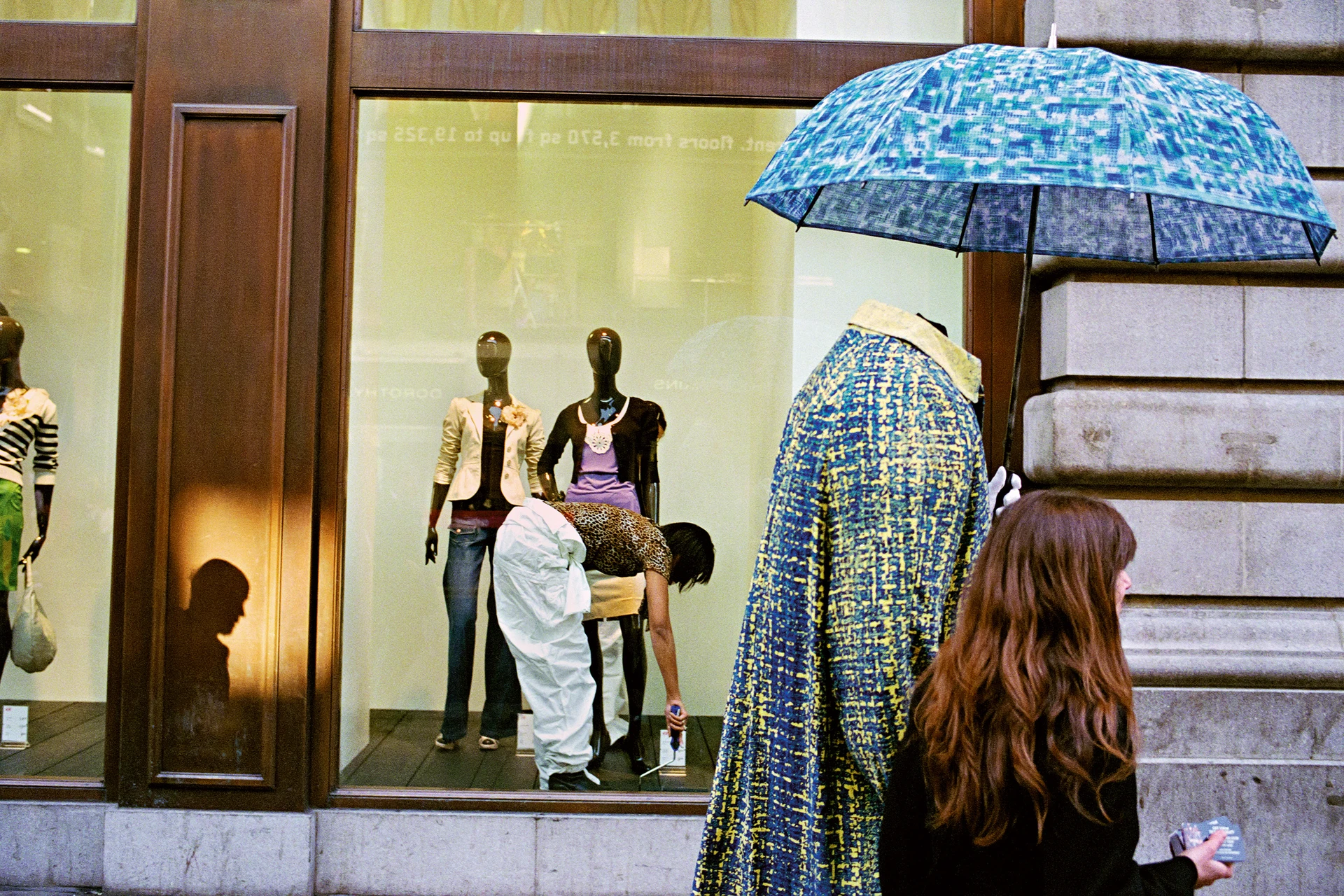 A view towards a shop window showing several mannequins. On the left, the silhouette of a man is seen as a shadow in a reflection of the sun. In the centre, a woman is bent over, working behind the window. To the right, two people with umbrellas pass by on the pavement – one of whom appears to be missing their head.