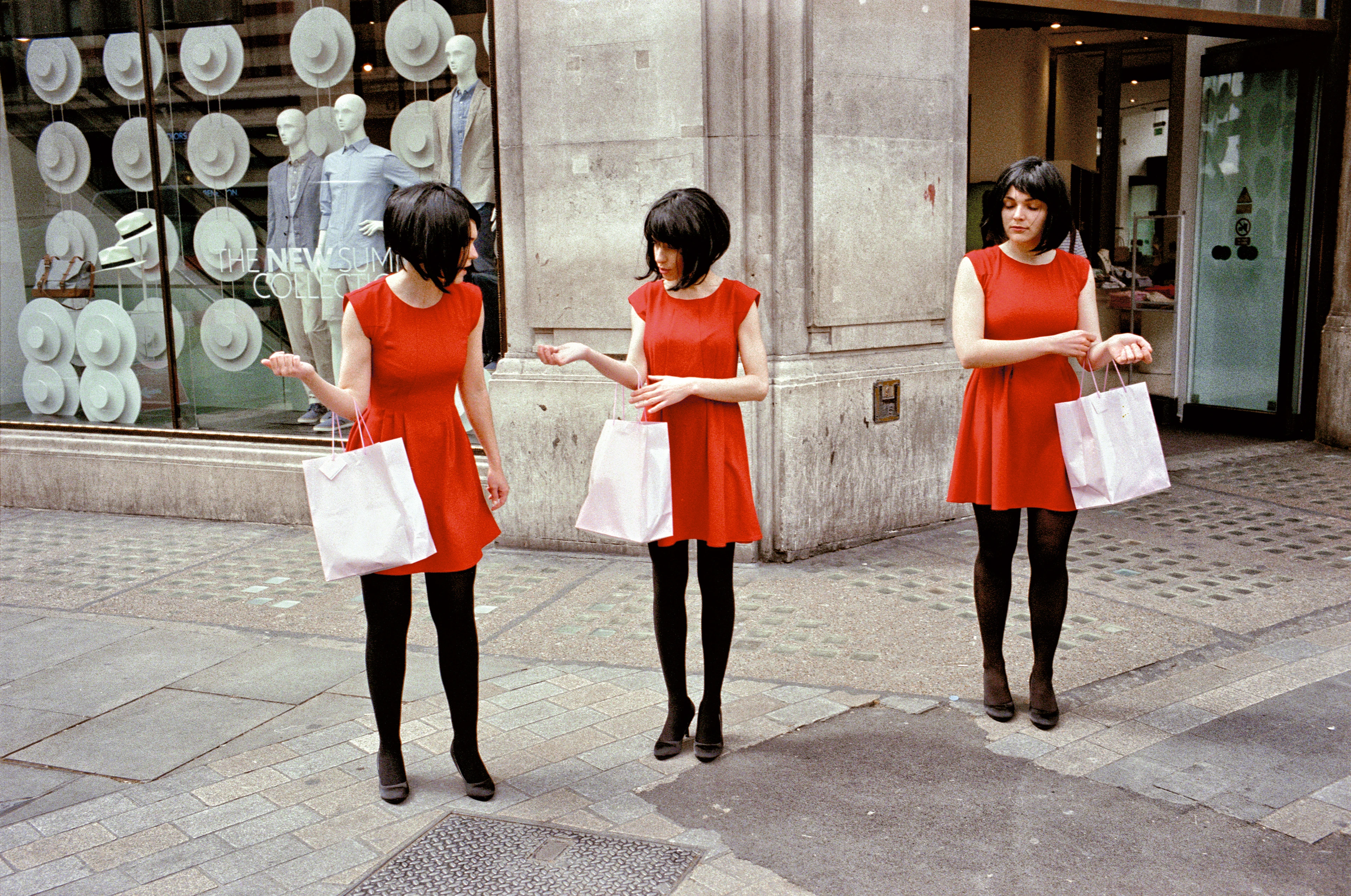 Three women wearing identical red dresses, wigs and black tights stand side by side on the pavement, each holding a white shopping bag.