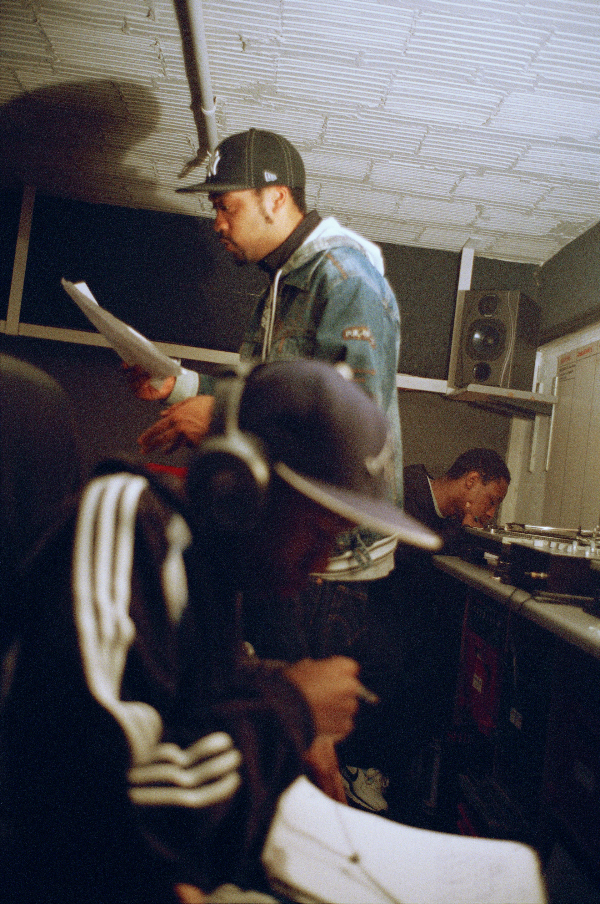 Trzech mężczyzn w piwnicy z gramofonami coś zapisuje, podczas gdy jeden z nich rapuje na stojąco.