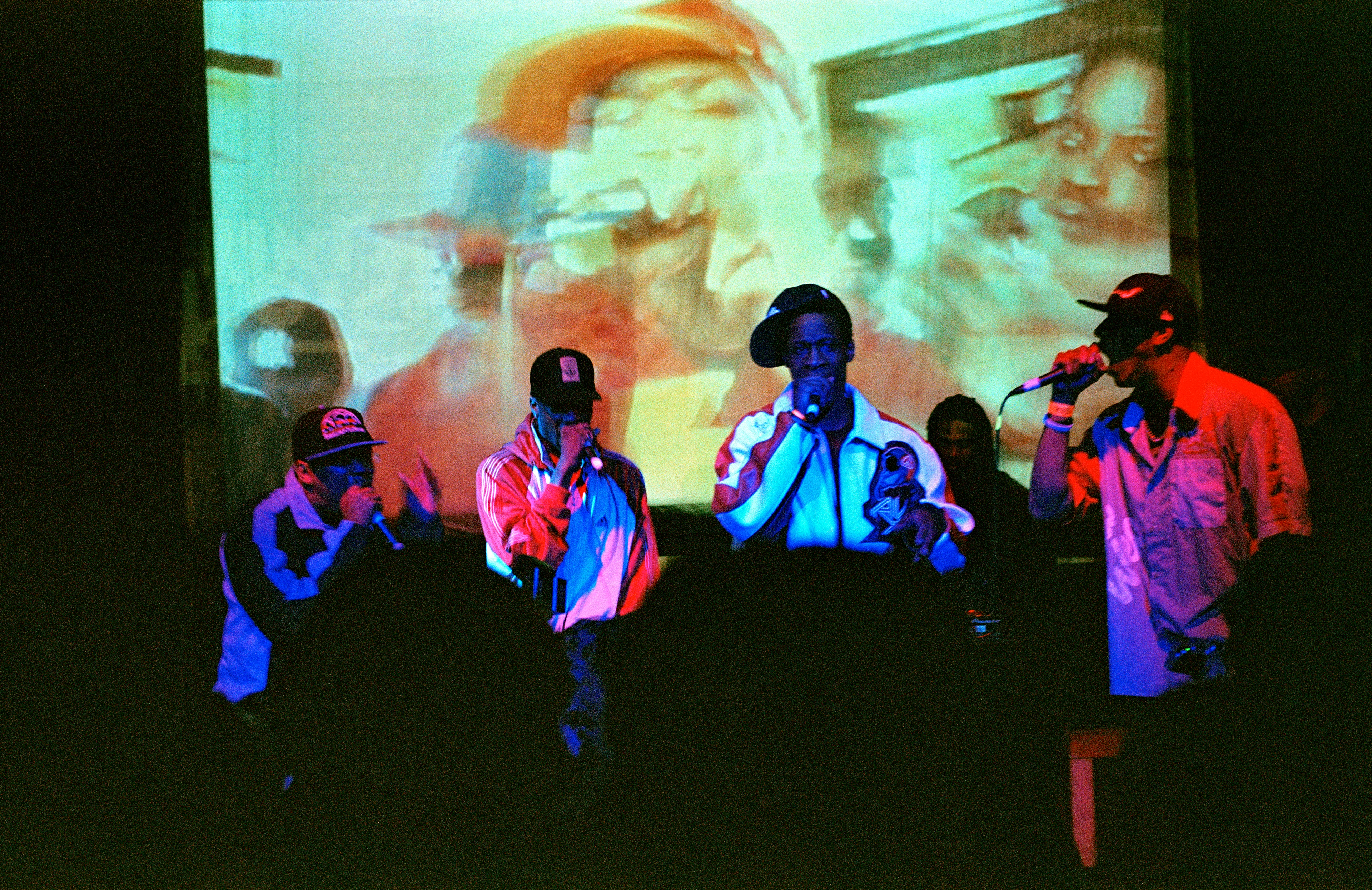 Kilku kolorowo oświetlonych, młodych, czarnych mężczyzn stoi na scenie z mikrofonami w rękach, a w tle widoczny jest obraz z projektora.