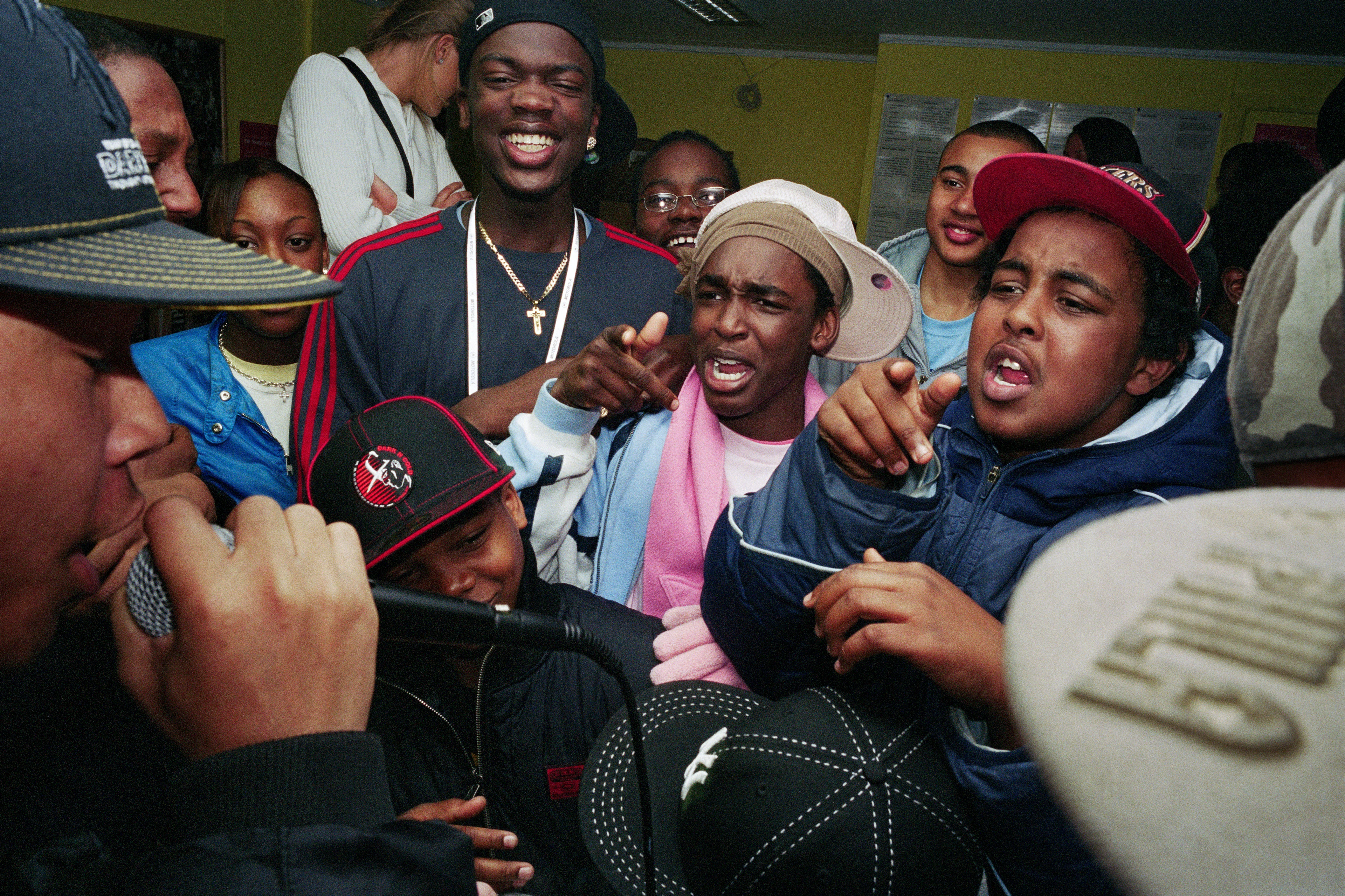 Młodzi, czarni mężczyźni stają stłoczeni w pomieszczeniu i euforycznie dopingują rapera rapującego do mikrofonu na pierwszym planie.