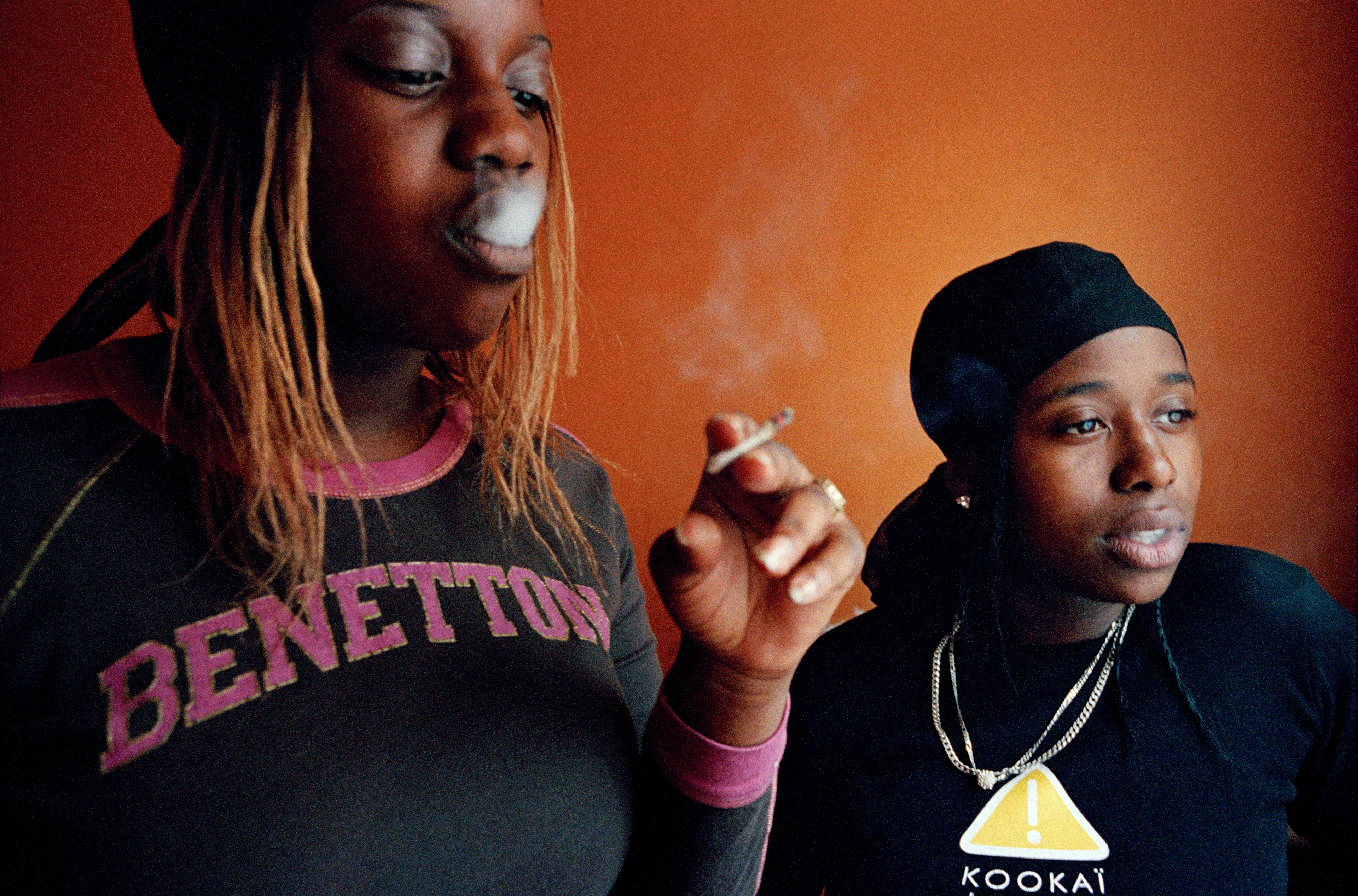 Portret dwóch młodych, czarnych kobiet w strojach ulicznych, z których jedna bawi się dymem papierosa, obie patrzą z zamyśleniem w stronę przeciwną do aparatu.