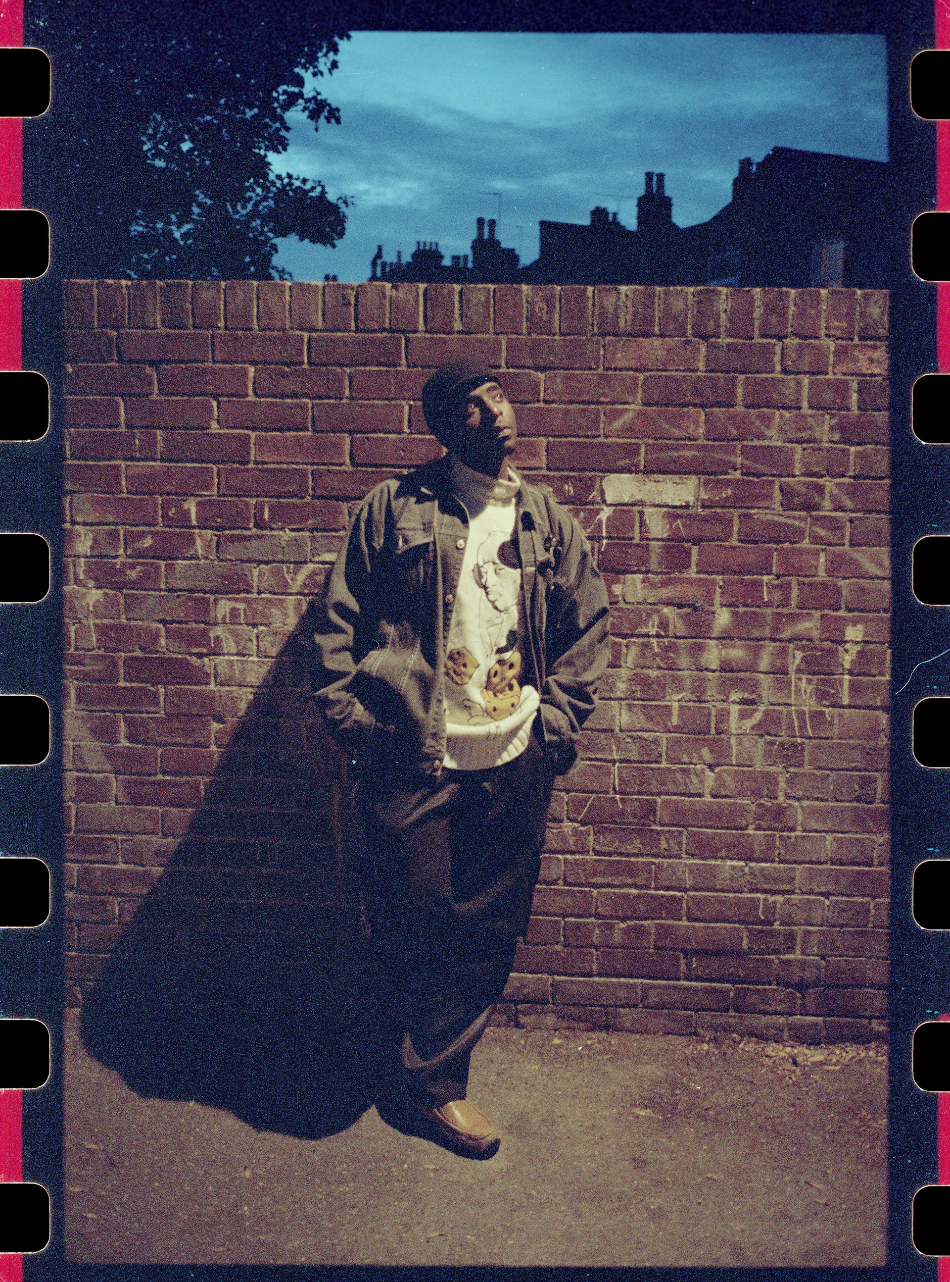 Porträt des Schwarzen jungen TUGGSTAR der in Streetwear in der Dämmerung lässig an einer Mauer posiert und in Richtung Himmel schaut.