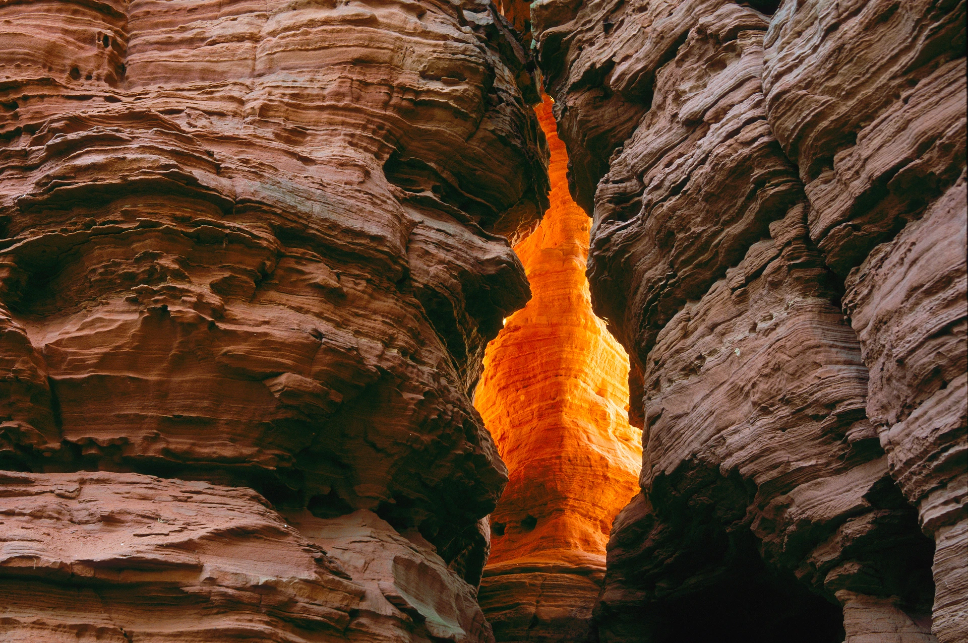 Roccia di arenaria rossa con una stretta fessura attraverso la quale filtra la luce calda del sole, che tinge le rocce di un arancione brillante.