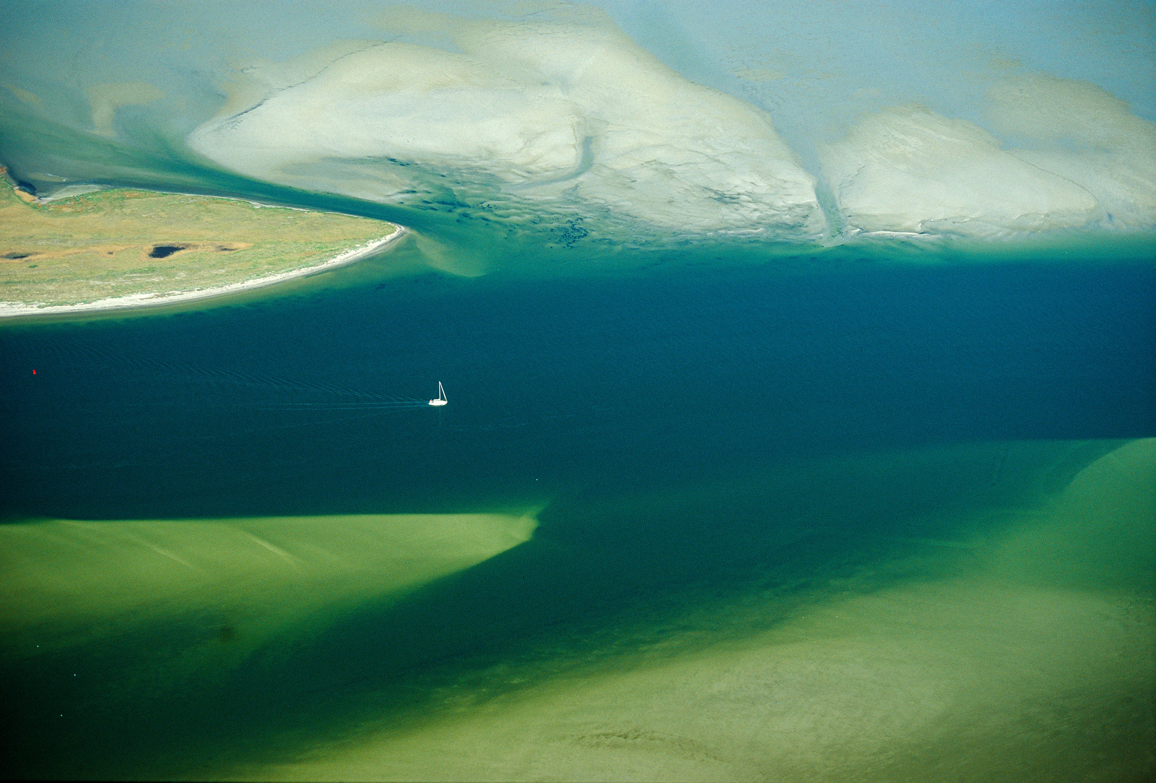 Una barca a vela bianca fotografata da una vista aerea, che veleggia solitaria su un canale verde-blu dall’effetto pittorico tra isole e banchi di sabbia.