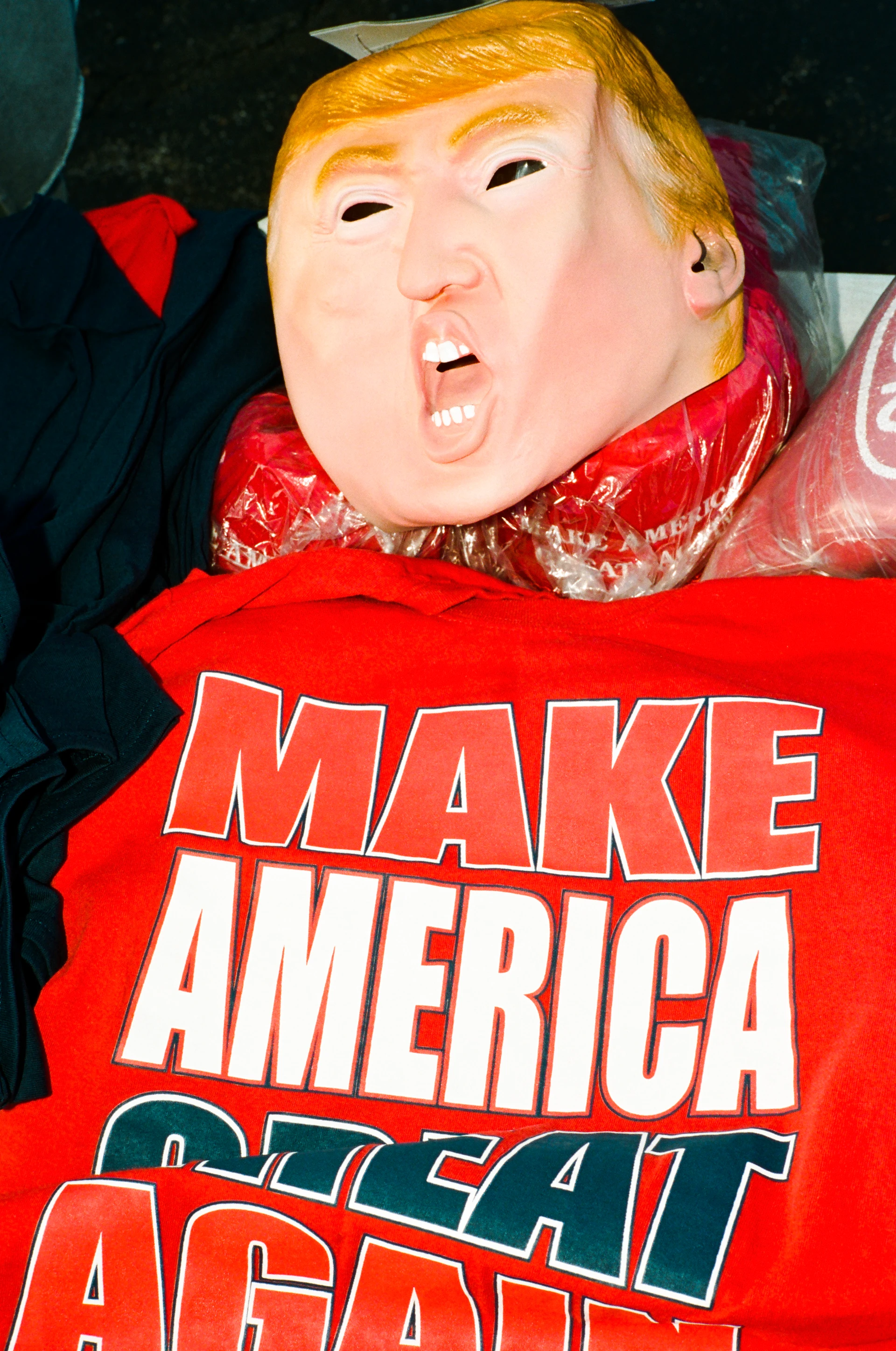 A rubber mask of Donald Trump lies atop other merchandise, seemingly wearing a red T-shirt bearing the slogan Make America Great Again.