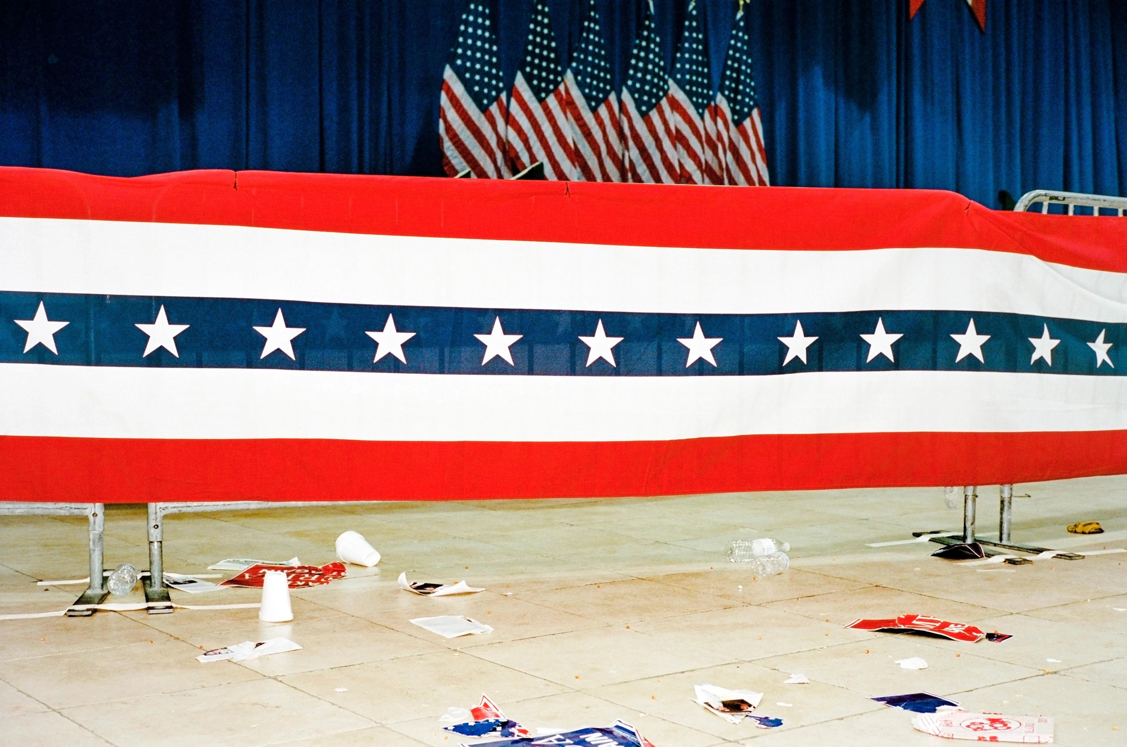 An empty hall with red, white and blue banners pinned to the stage and US flags in the background. Scattered paper cups, rubbish and remnants of flags are on the floor in front of it.