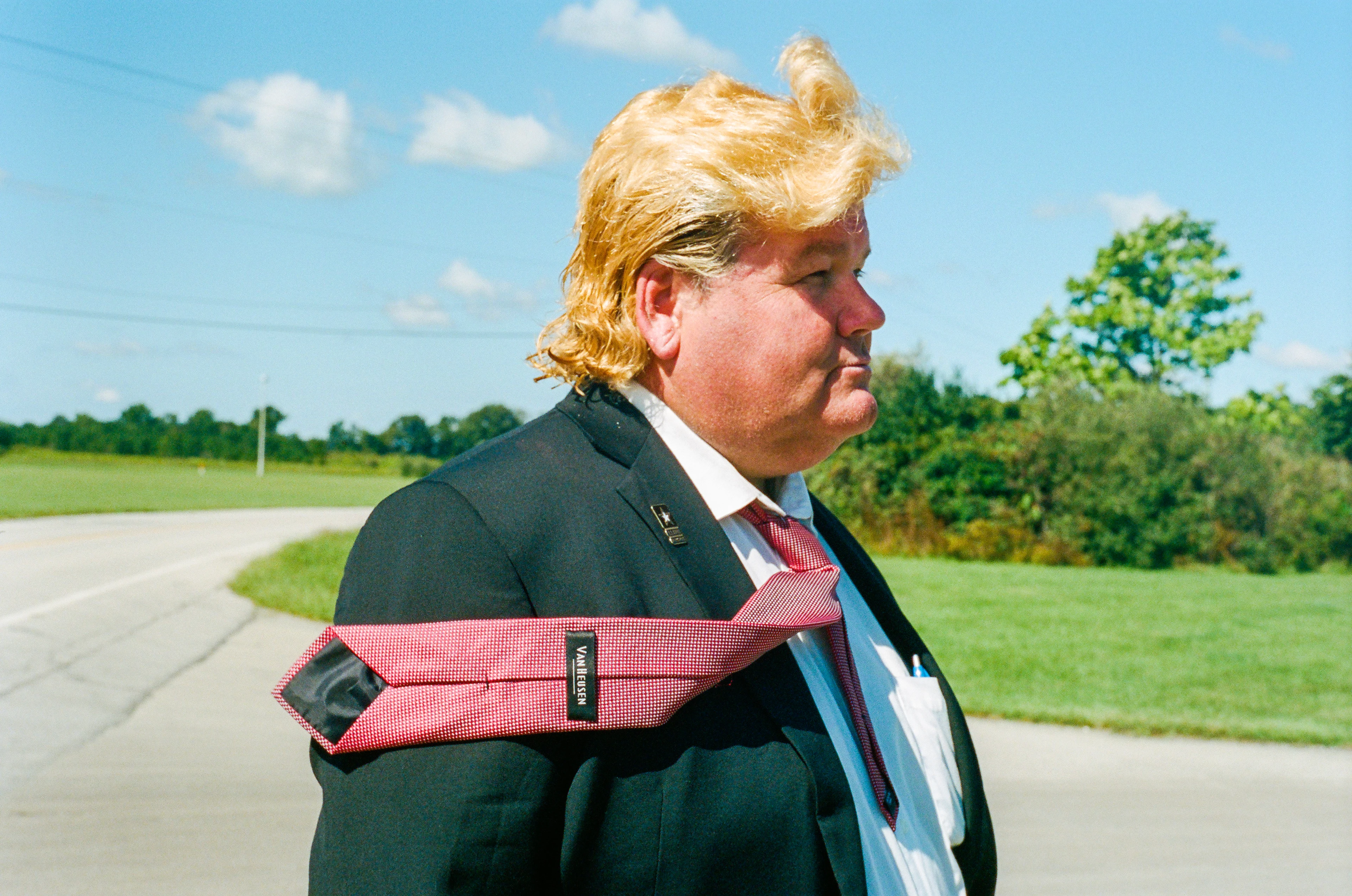 A man in a business suit wearing a Donald Trump-style wig and tie stands on the side of the road in front of a green area and blue sky.