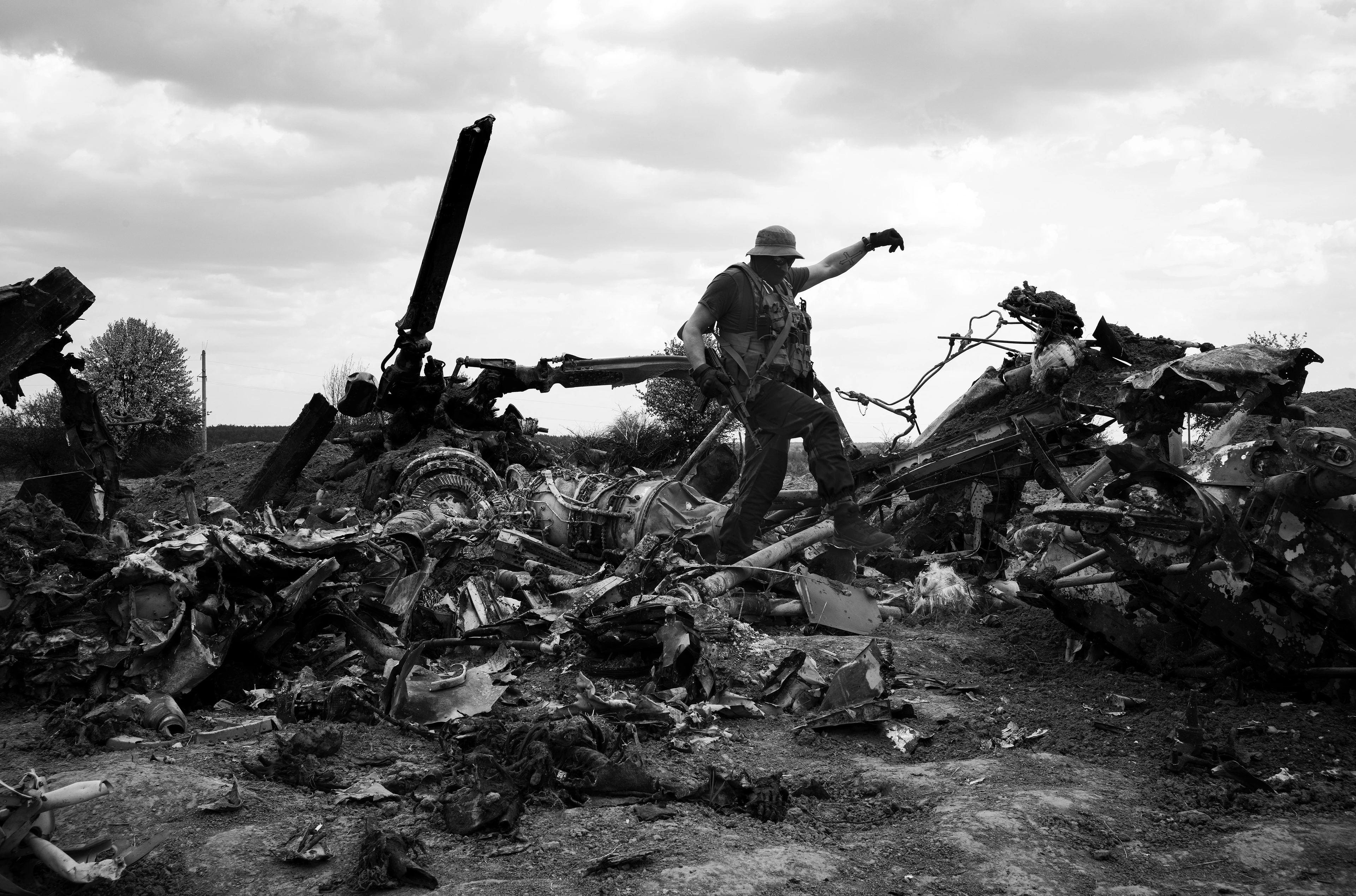 Schwarz-Weiß-Bild: Ein Mann in Militärkleidung klettert über die verdrehten, verkohlten Überreste eines zerstörten Hubschraubers, die über ein Feld verstreut liegen.
