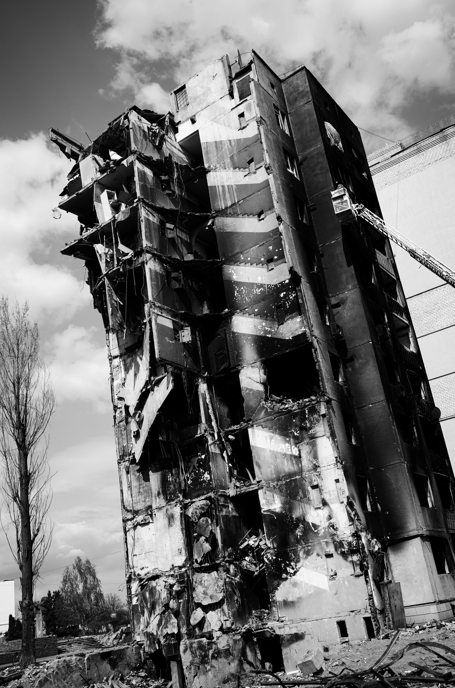 Schwarz-Weiß-Bild: Ein mehrstöckiges Gebäude steht frei auf einem Platz, stark zerbombt mit aufgerissenen Etagen und herabhängenden Betonplatten, ein Krankorb fährt an eins der oberen Stockwerke heran.