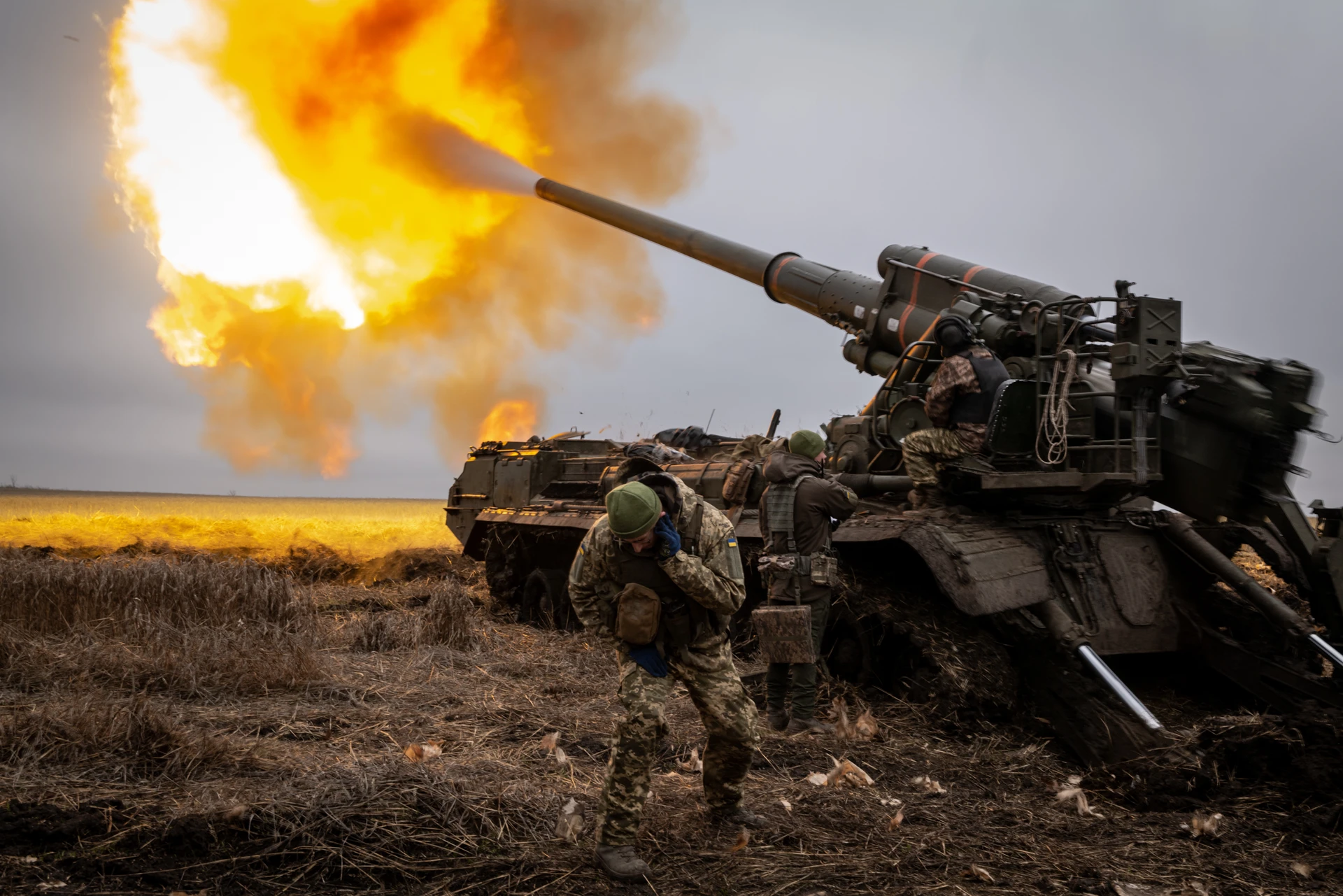 Ukrainian soldiers fire a large artillery cannon in an open field, creating a bright fireball, while a soldier ducks for cover.