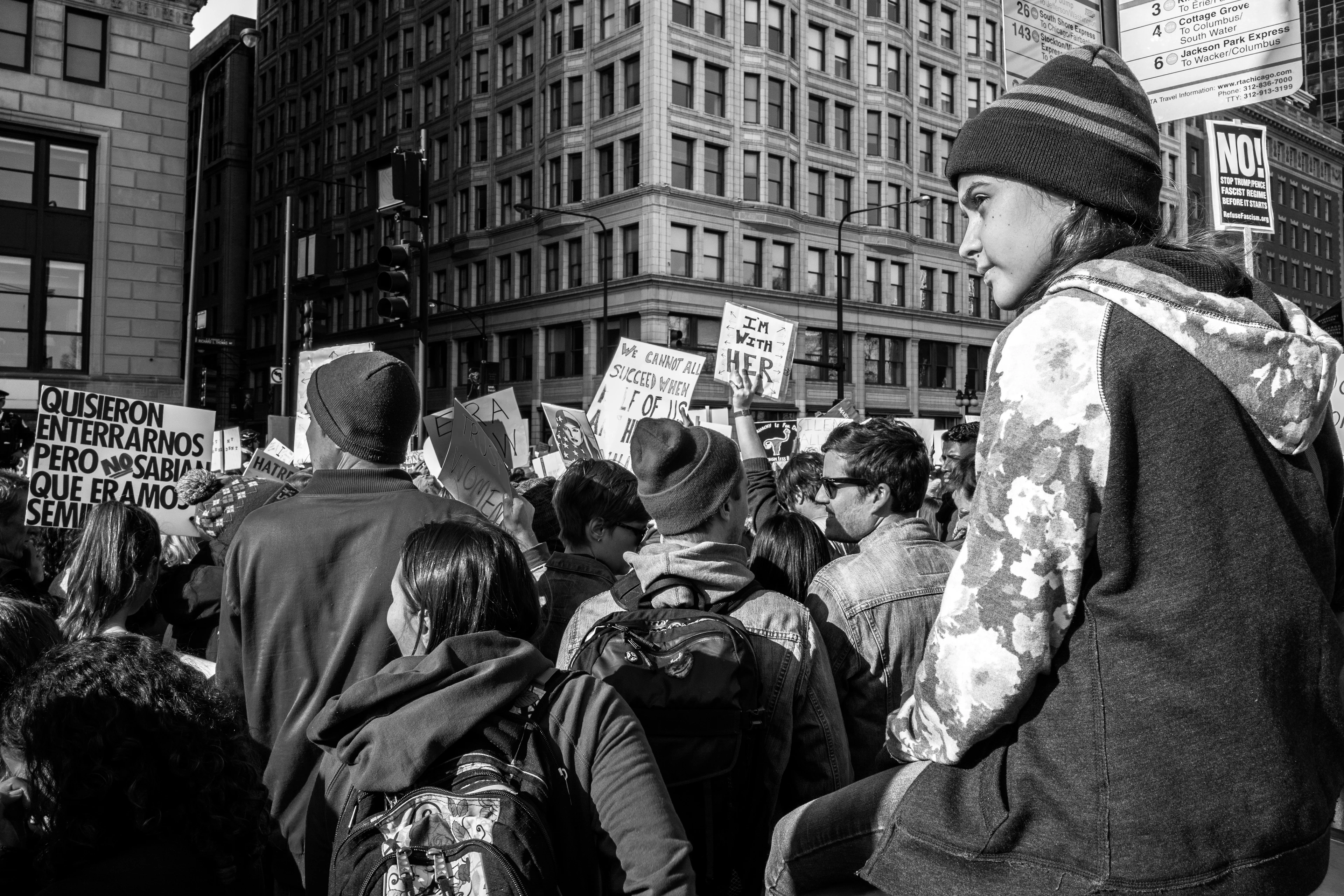 Black-and-white image: A girl sits elevated on the edge of a Women’s March demonstration, attentively observing the events in front of her amidst an urban environment and a crowd of people holding placards.