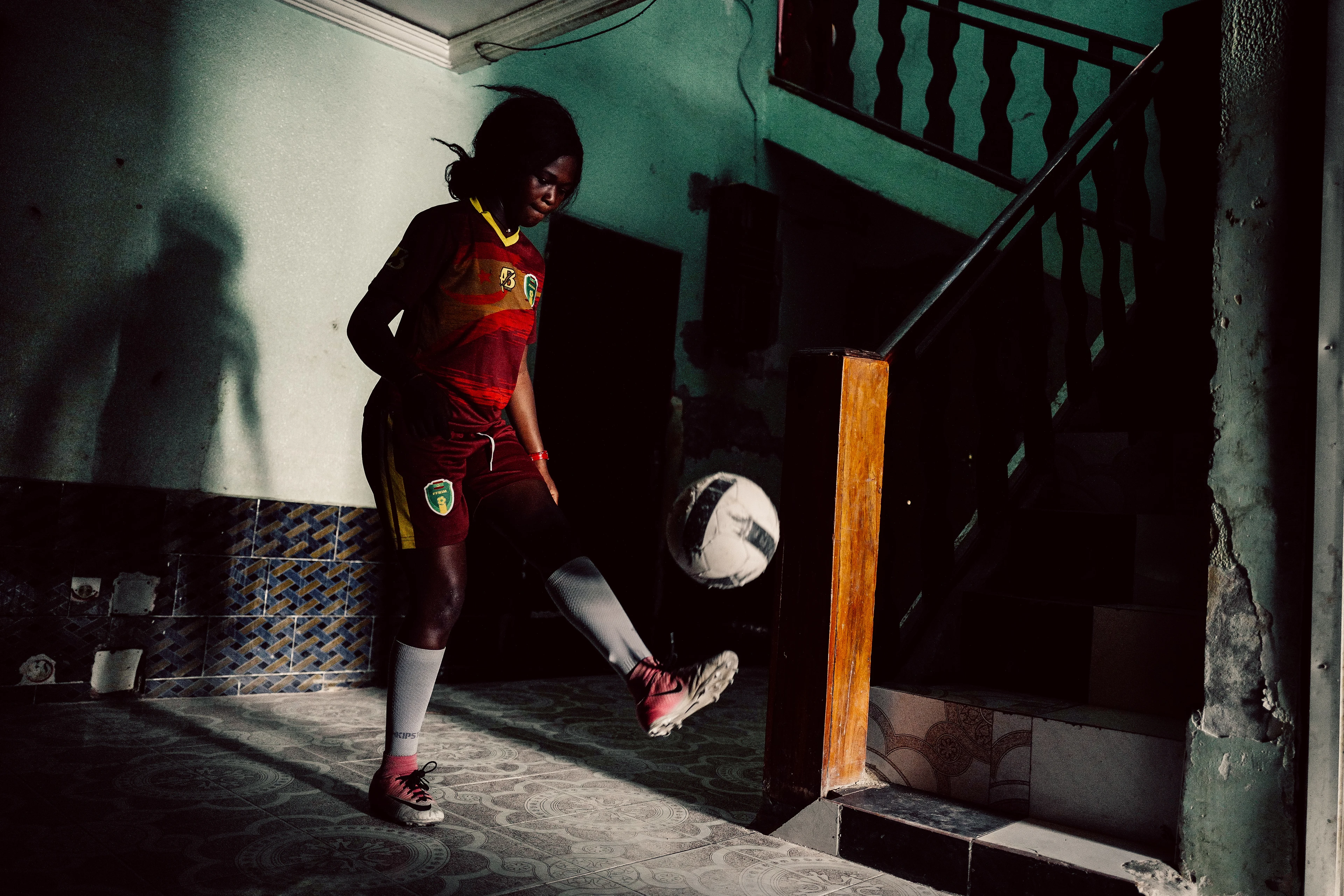 A woman wearing a full football kit plays with a ball in a stairwell, kicking it into the air with her foot, while her shadow is projected onto the wall.