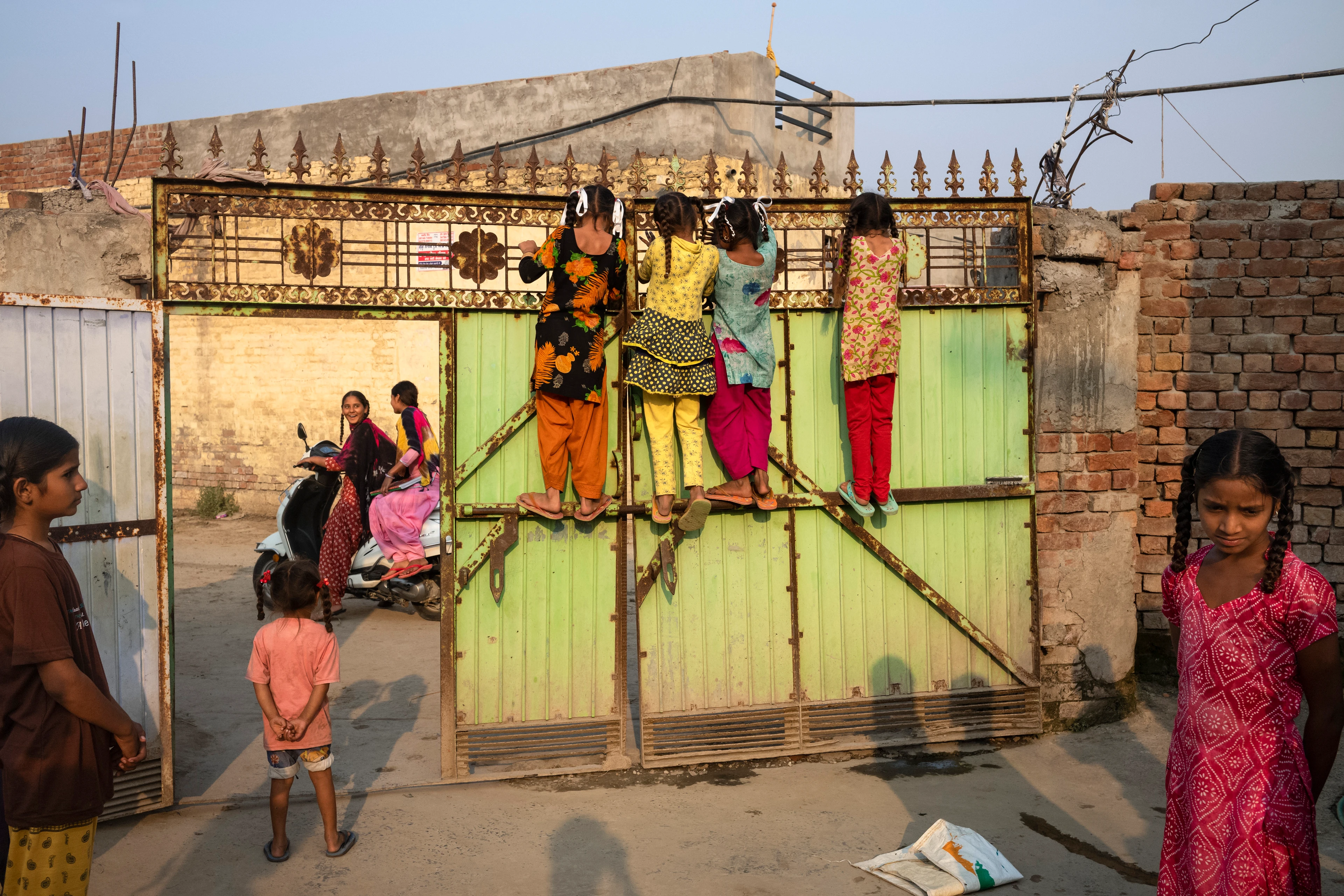 Four girls dressed in Indian clothes stand on a metal gate and look over it at the street, where two cheerful young women sit on a scooter and drive past. Other girls stand in the courtyard and watch.