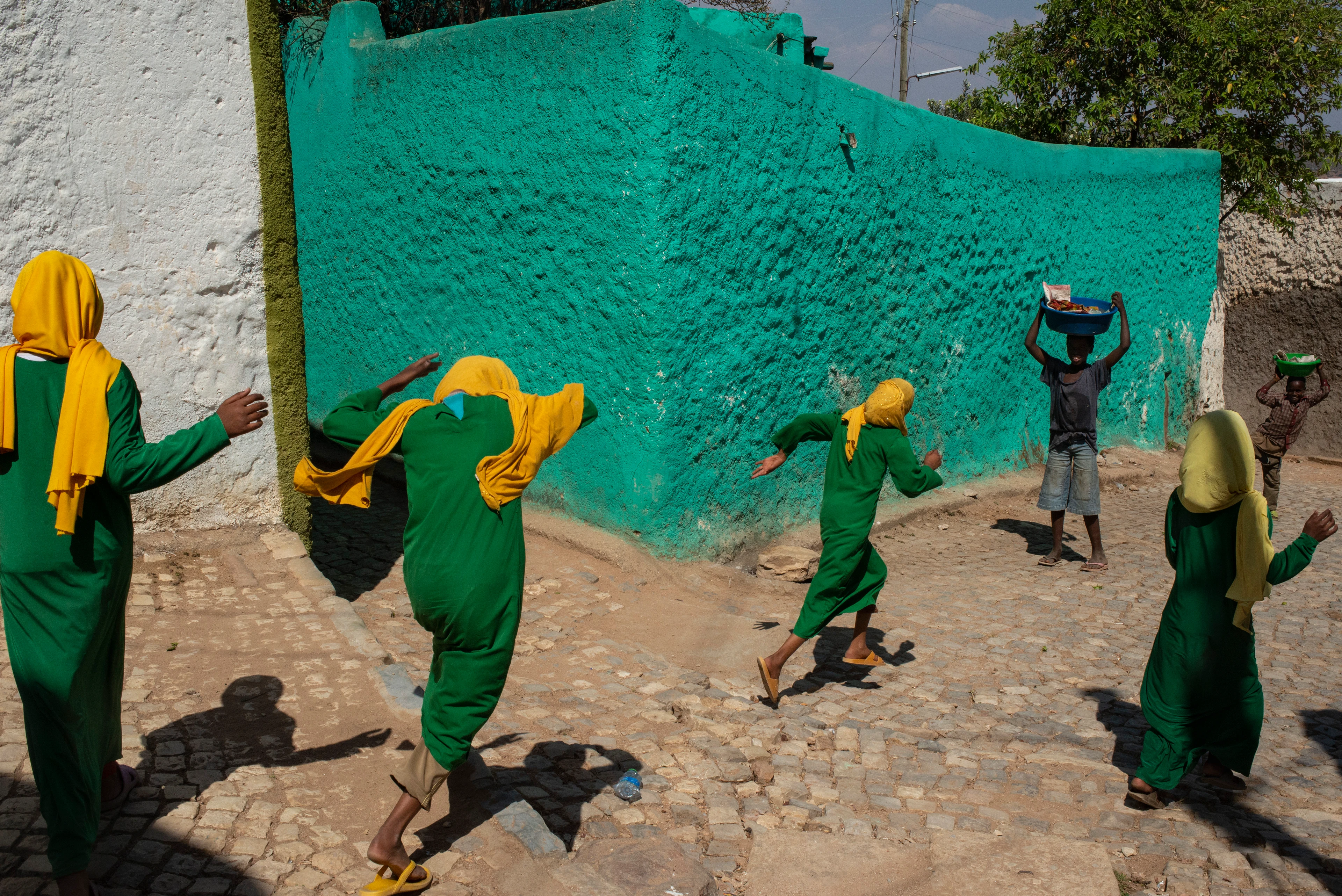 A group of Black girls wearing green tunics and yellow headscarves run along a cobbled path in front of a turquoise wall, playing with two Black boys.
