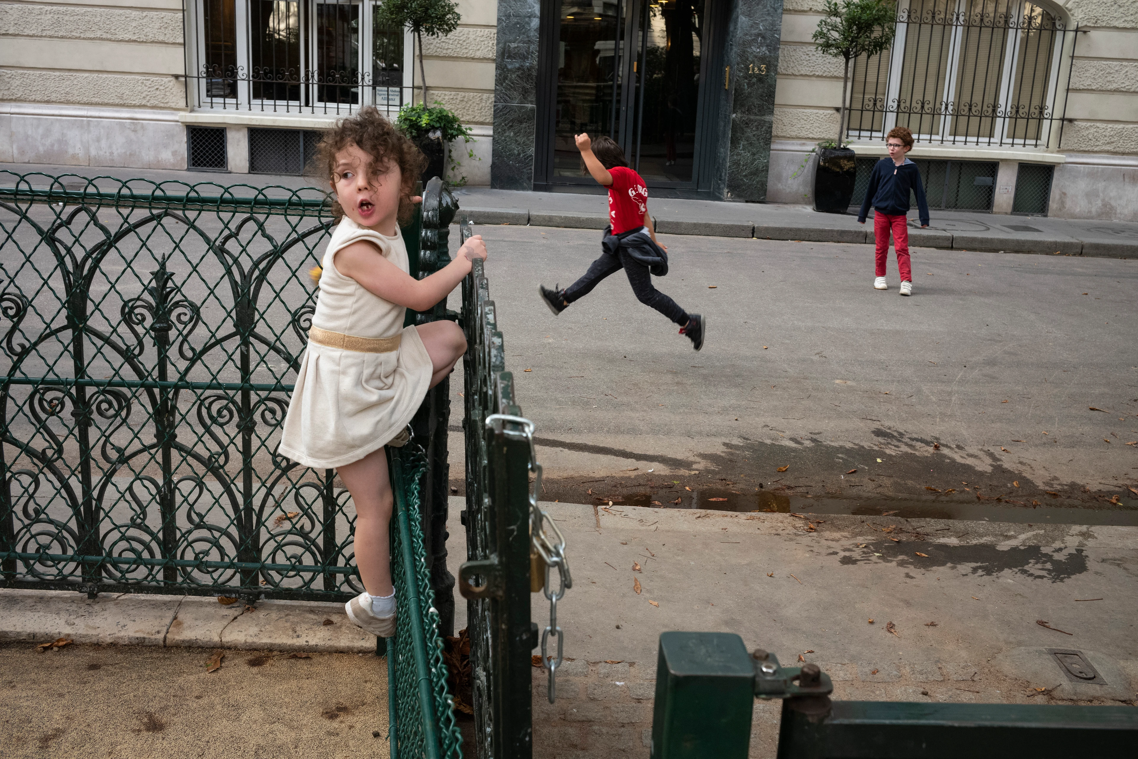 A girl wearing a light-coloured summer dress climbs up a metal fence, looking cheeky, while two other children run and jump around in the street behind her.