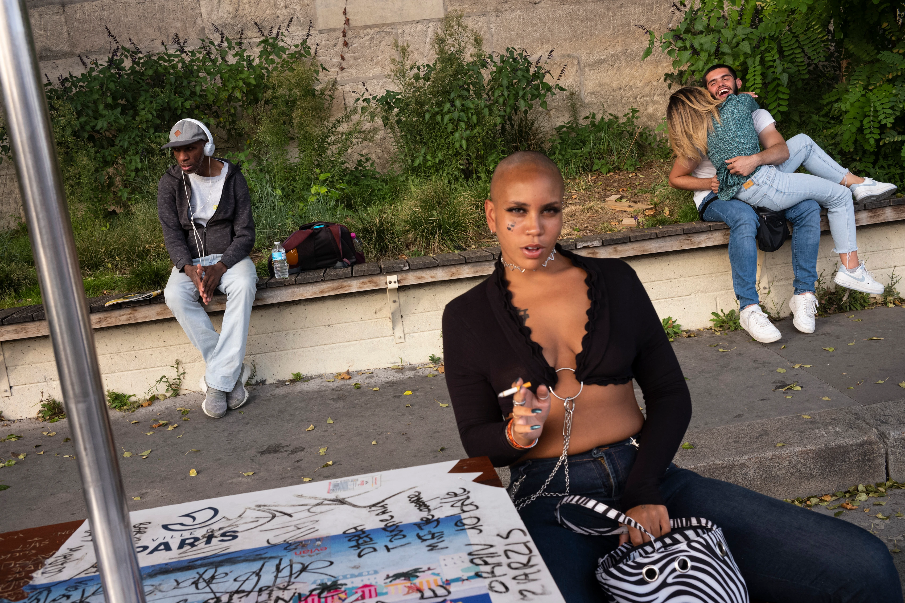 A woman with a shaved head sits confidently at a graffiti-covered table, smoking and looking at the camera. In the background, a couple hug tightly on a bench, while a boy with headphones sits alone nearby.