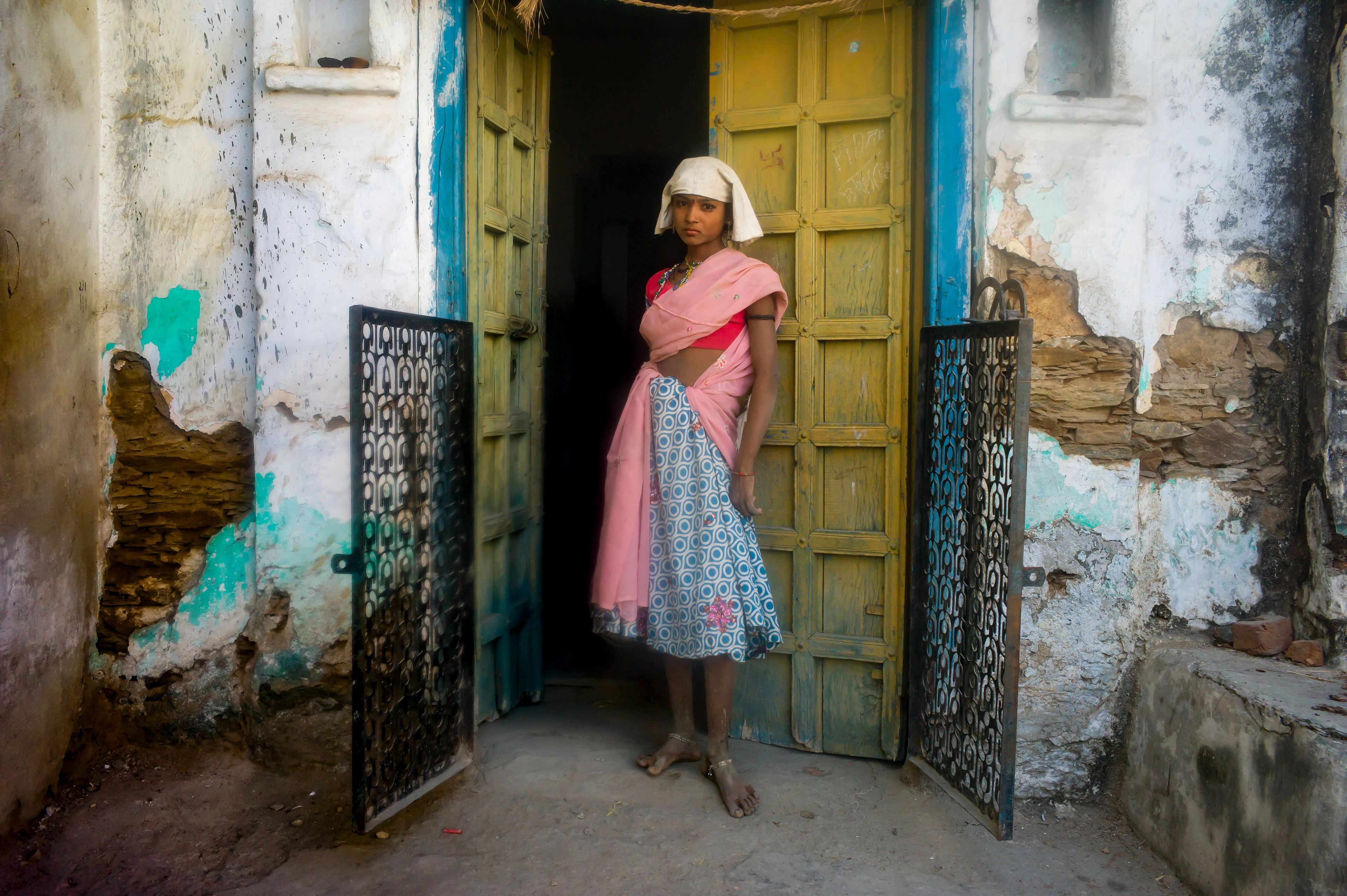 A young woman with a dark complexion, wearing traditionally colourful clothes, stands barefoot in an open doorway with weathered walls. She has a painted dot between her eyes and looks confidently at the camera.