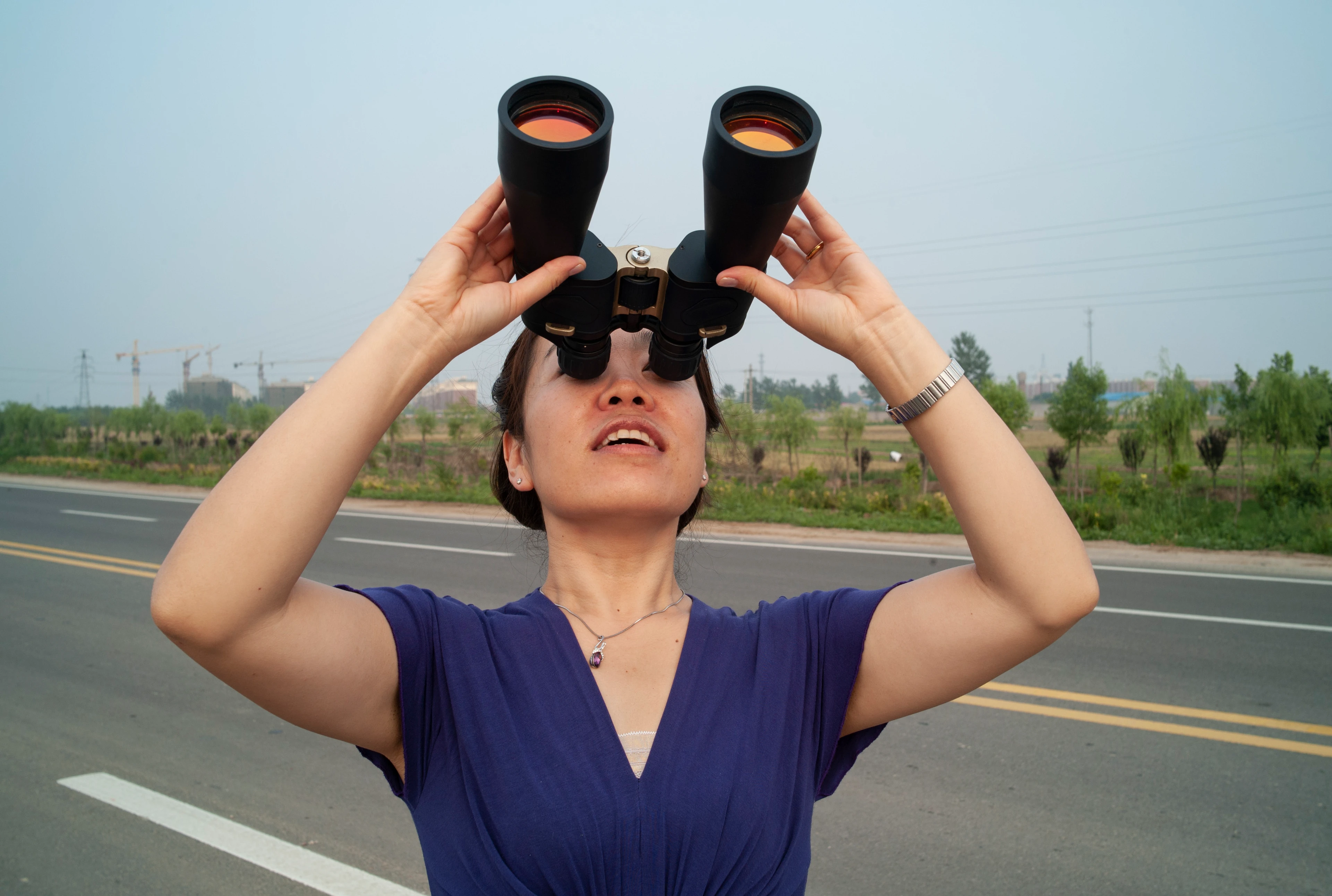 An Asian woman stands on a country highway, holding a large pair of binoculars to her eyes, smiling as she looks up at the sky.