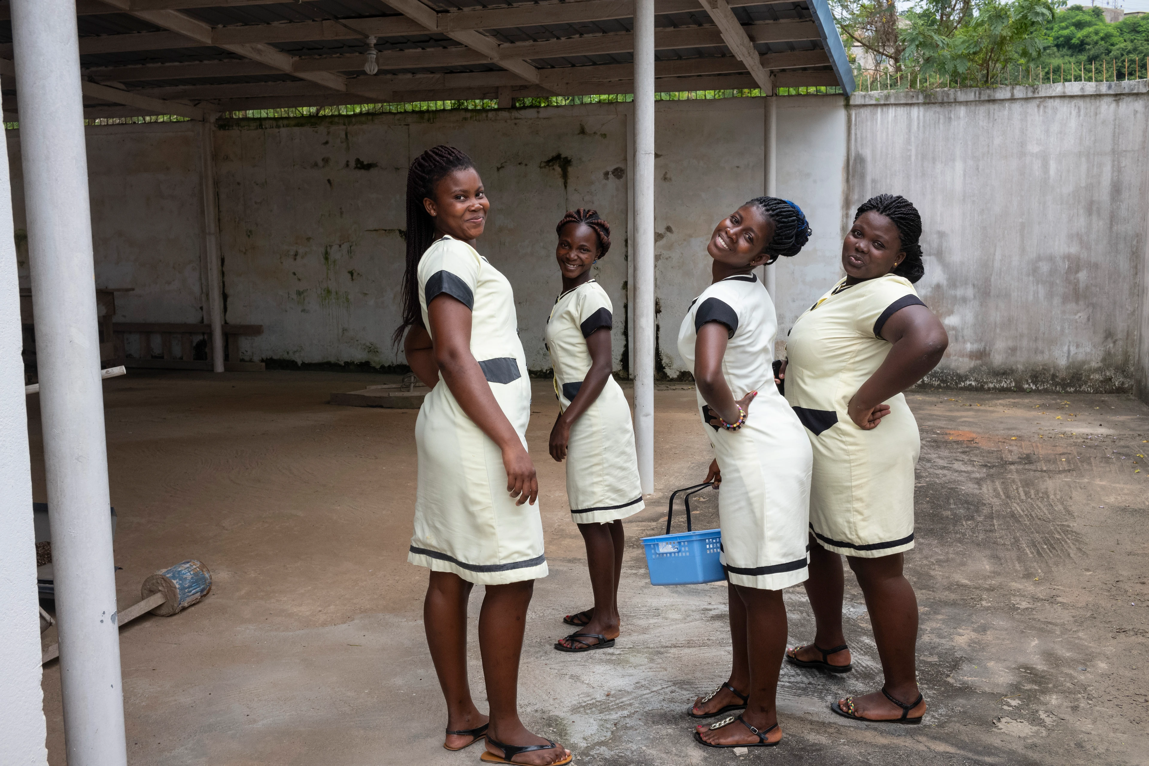 Four African women wearing the same white dress pose for the camera in a covered courtyard, smiling happily.