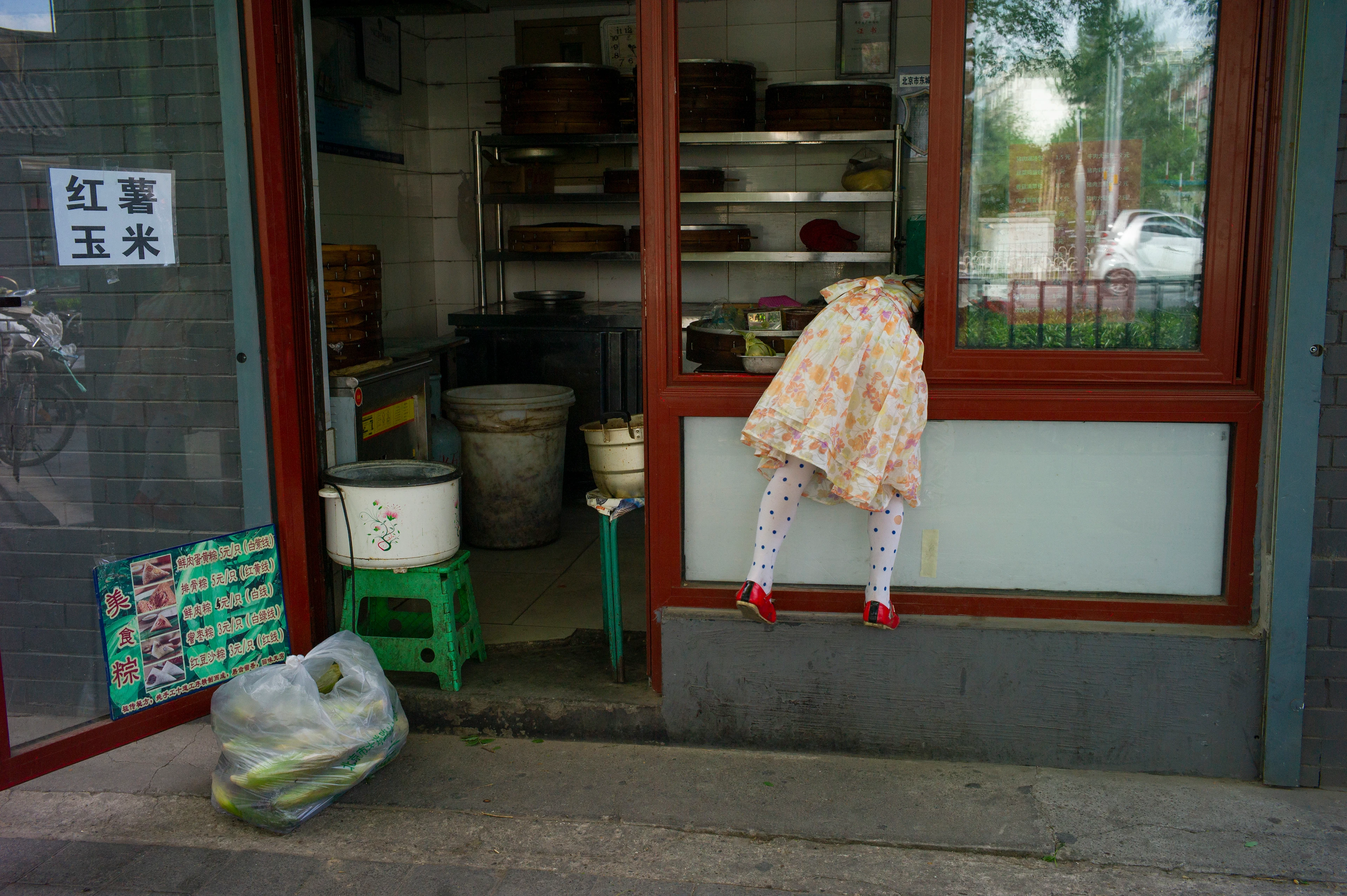 A child climbs through the open window of a small Chinese restaurant kitchen. The door next to it is open, with a bag of vegetables lying in front of the entrance.