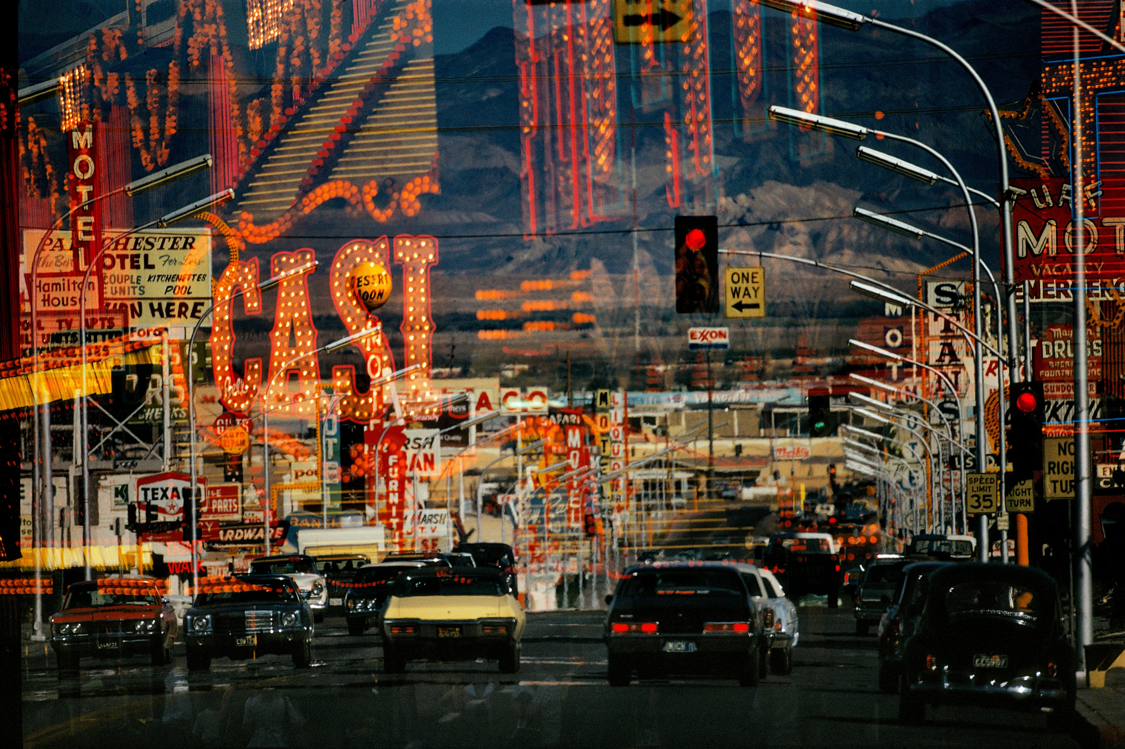 Una vista a través de una ventana a una calle con coches circulando y montañas al fondo en la que se reflejan las luces típicas de Las Vegas.