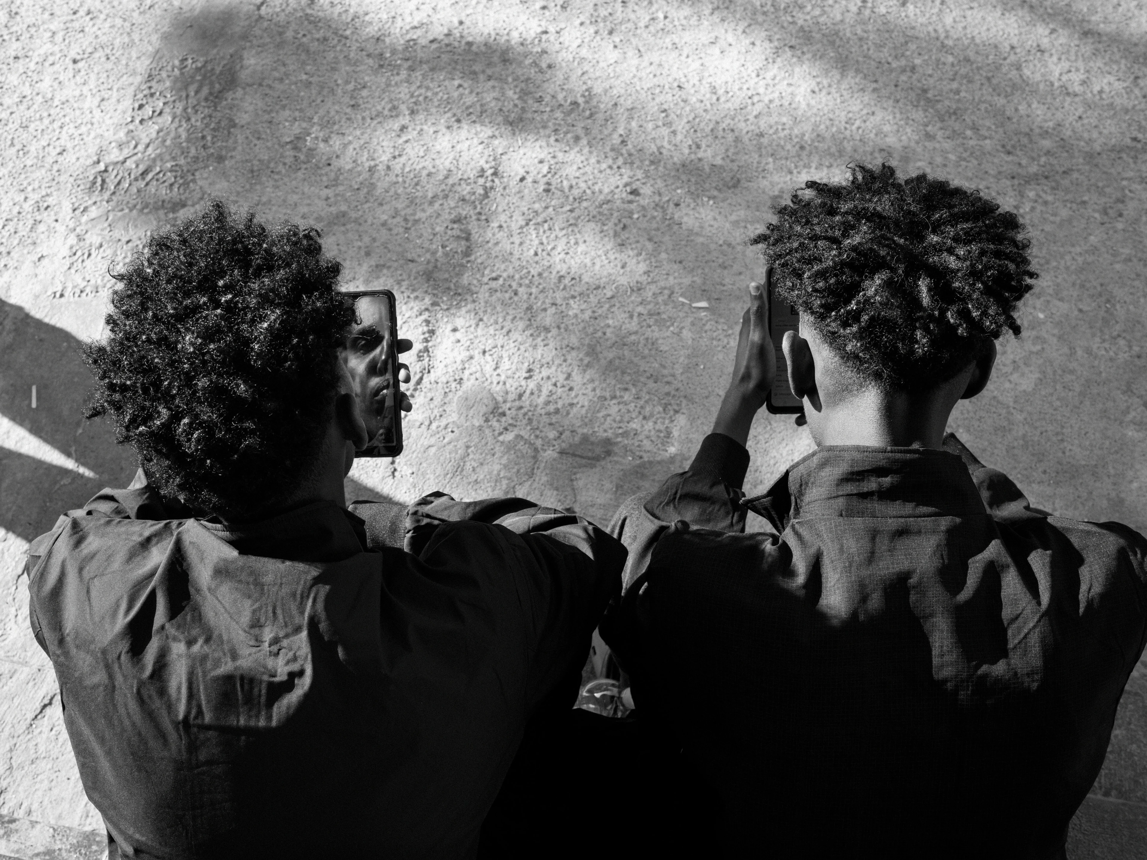 Black-and-white image: Two people sit next to each other with their backs to the camera, each looking at a smartphone on which the face of each person appears mirrored.