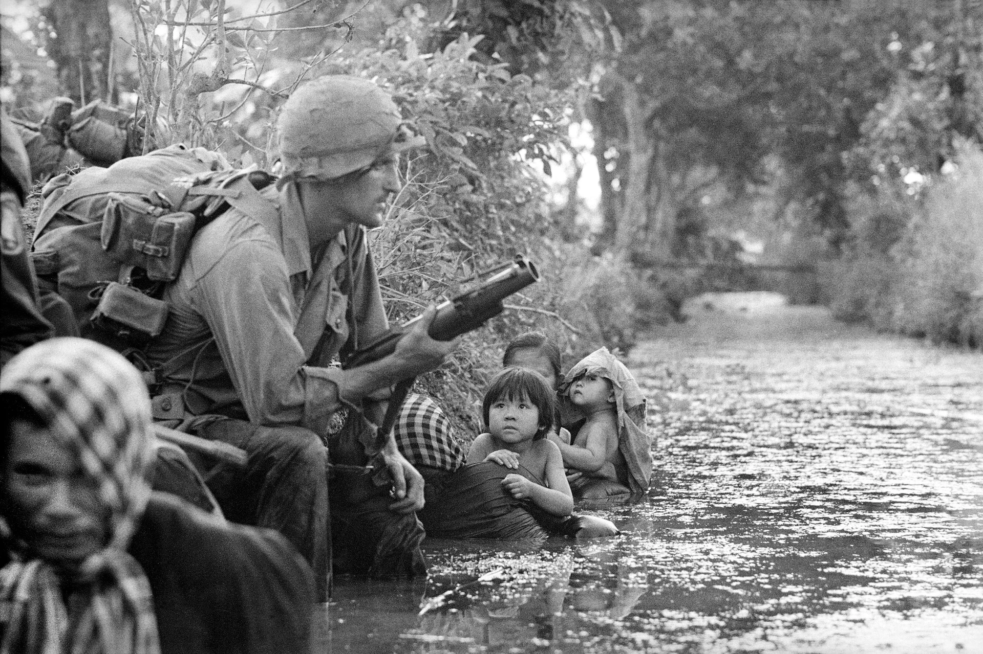 Motyw czarno-biały, wojna w Wietnamie, żołnierz klęczy nad rzeką, obserwowany przez wietnamskie dzieci
